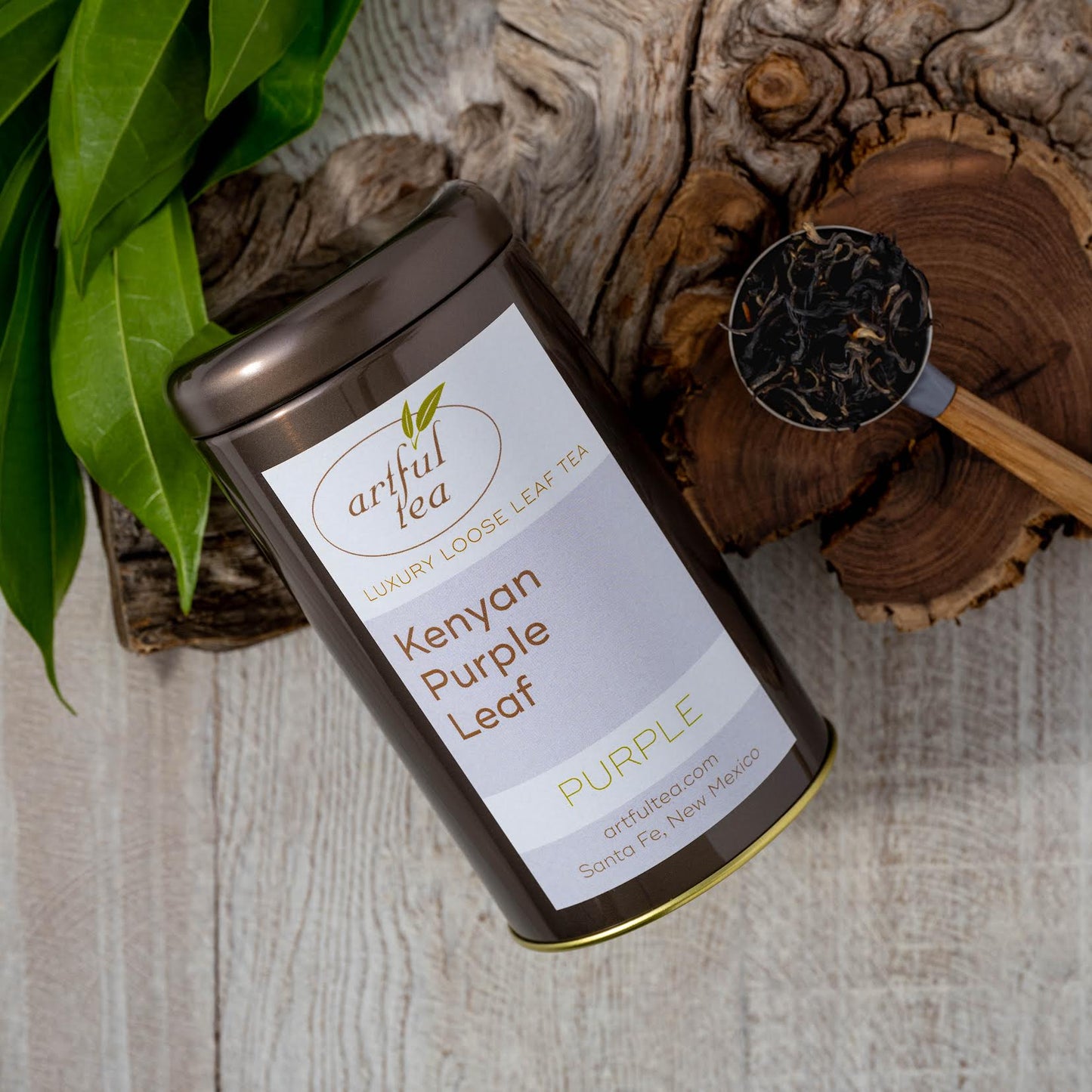 Kenyan Purple Leaf Tea shown packaged in a brown tin next to a wooden handled tablespoon full of loose tea leaves, both on top of a piece of wood.