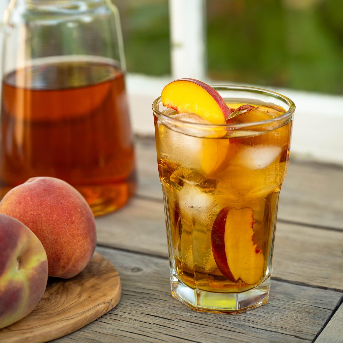 Ginger Peach Black Tea shown as iced tea with slices of a peach inside a clear cup on a wooden table. Another clear container full of tea and peaches nearby.