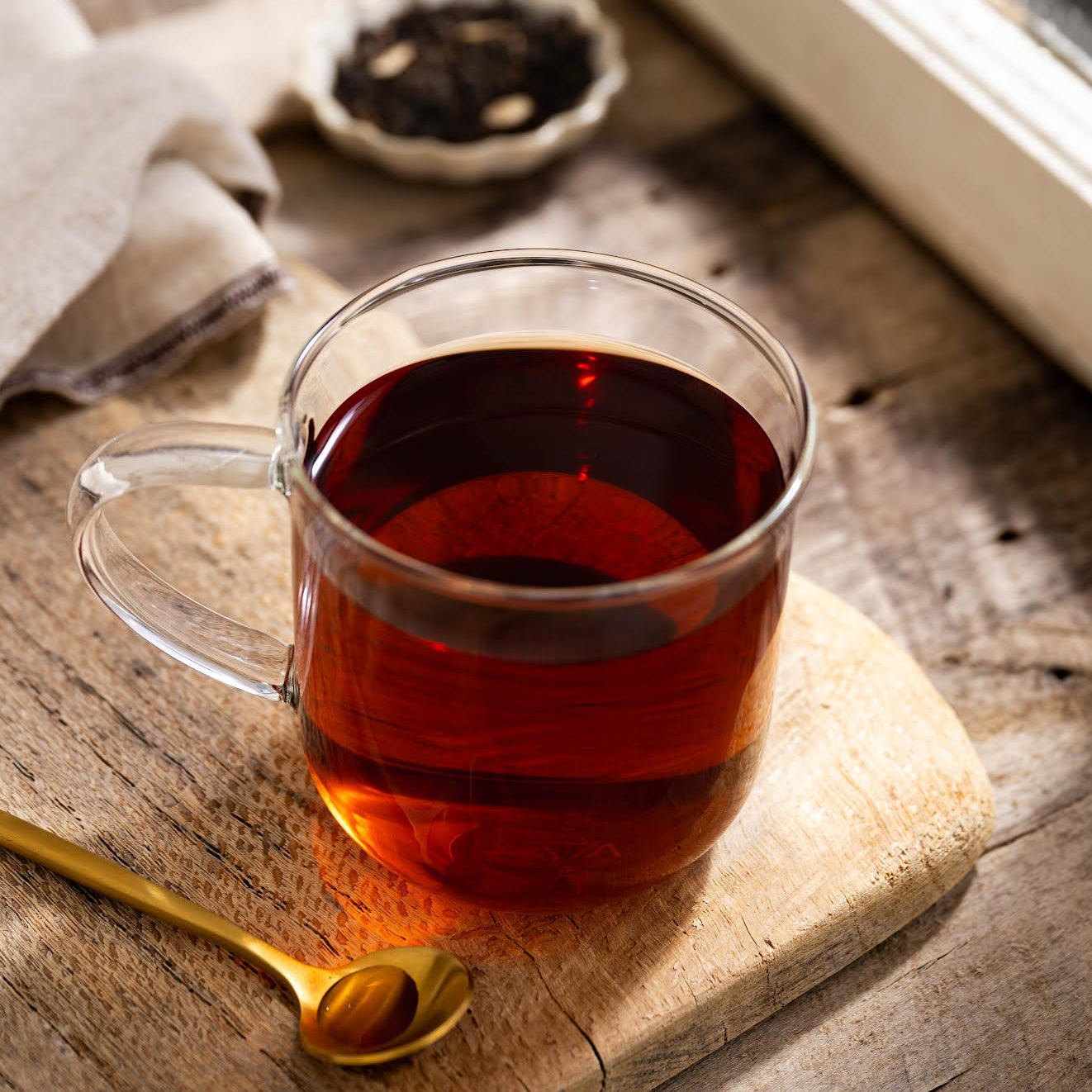 Caramel Pu-erh Tea shown as brewed tea in a clear mug on top of a wooden plank. Next to it is a golden spoon.