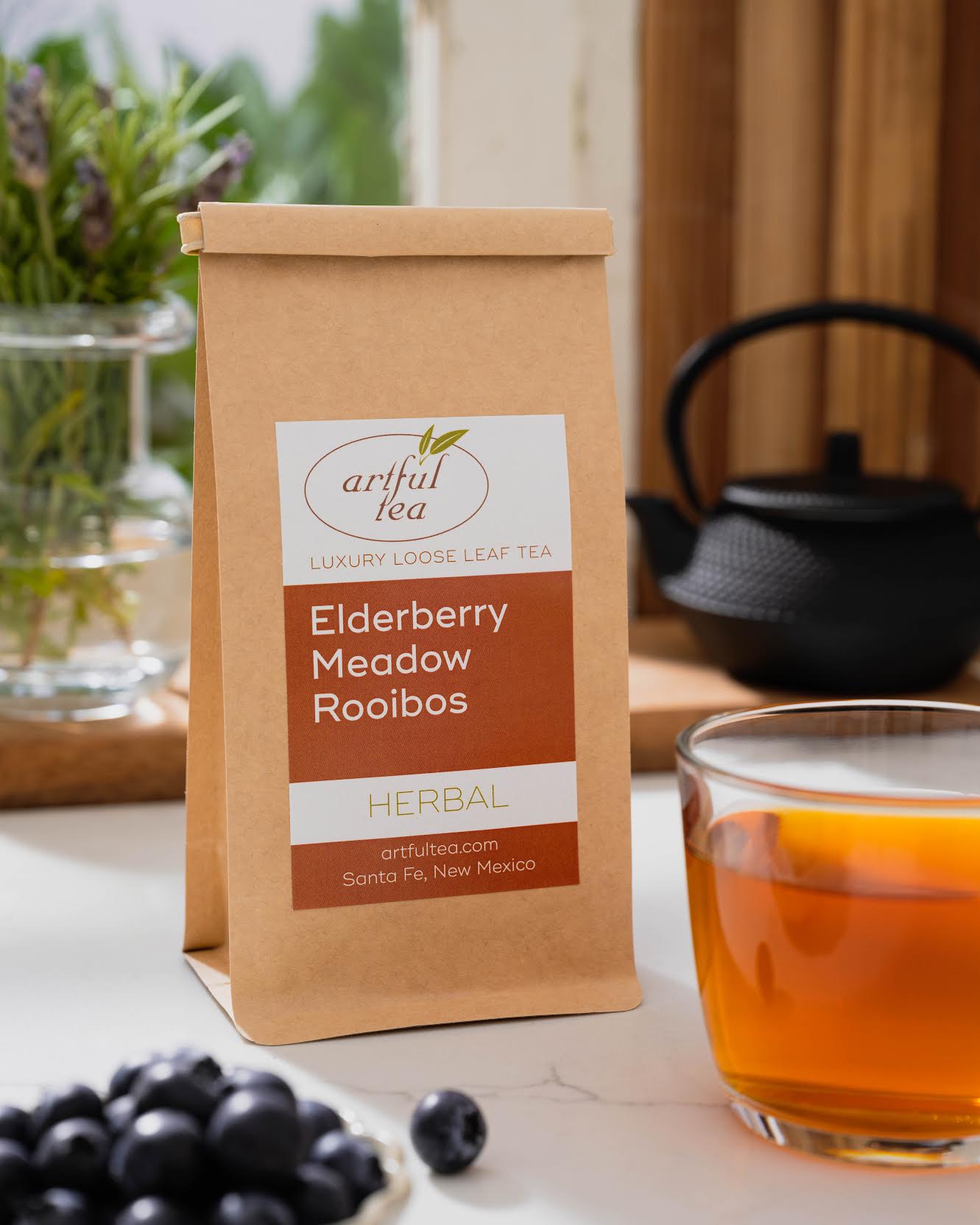 Elderberry Meadow Rooibos Herbal Tea shown packaged in a brown Kraft bag next to a cup of brewed tea and a small bowl of blueberries. In the background is a clear vase of flowers and a black teapot in a window sill.