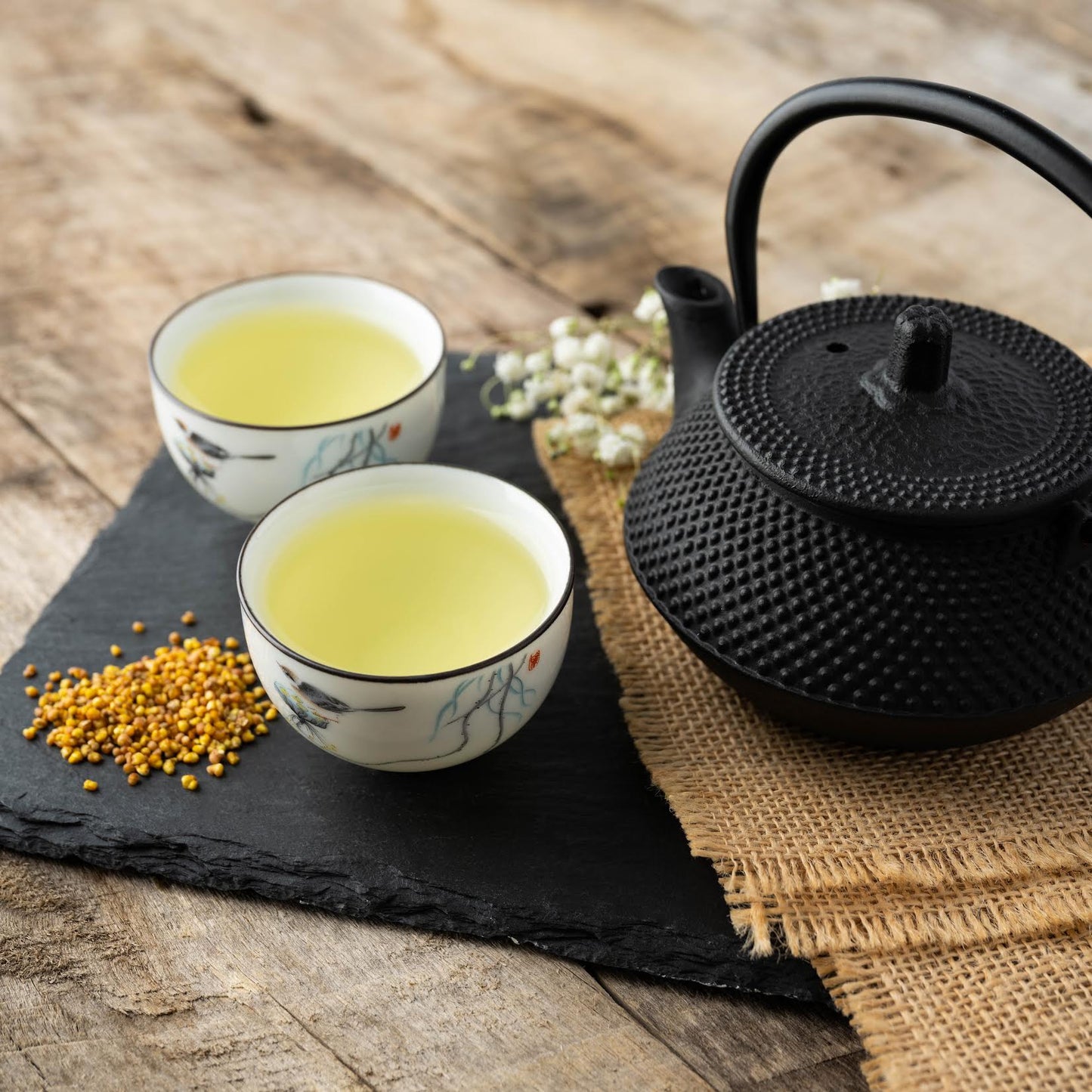 Sobacha Buckwheat Tea shown as two cups of brewed tea next to a black teapot on burlap, all on top of a black platter with loose buckwheat tea scattered next to the cups.