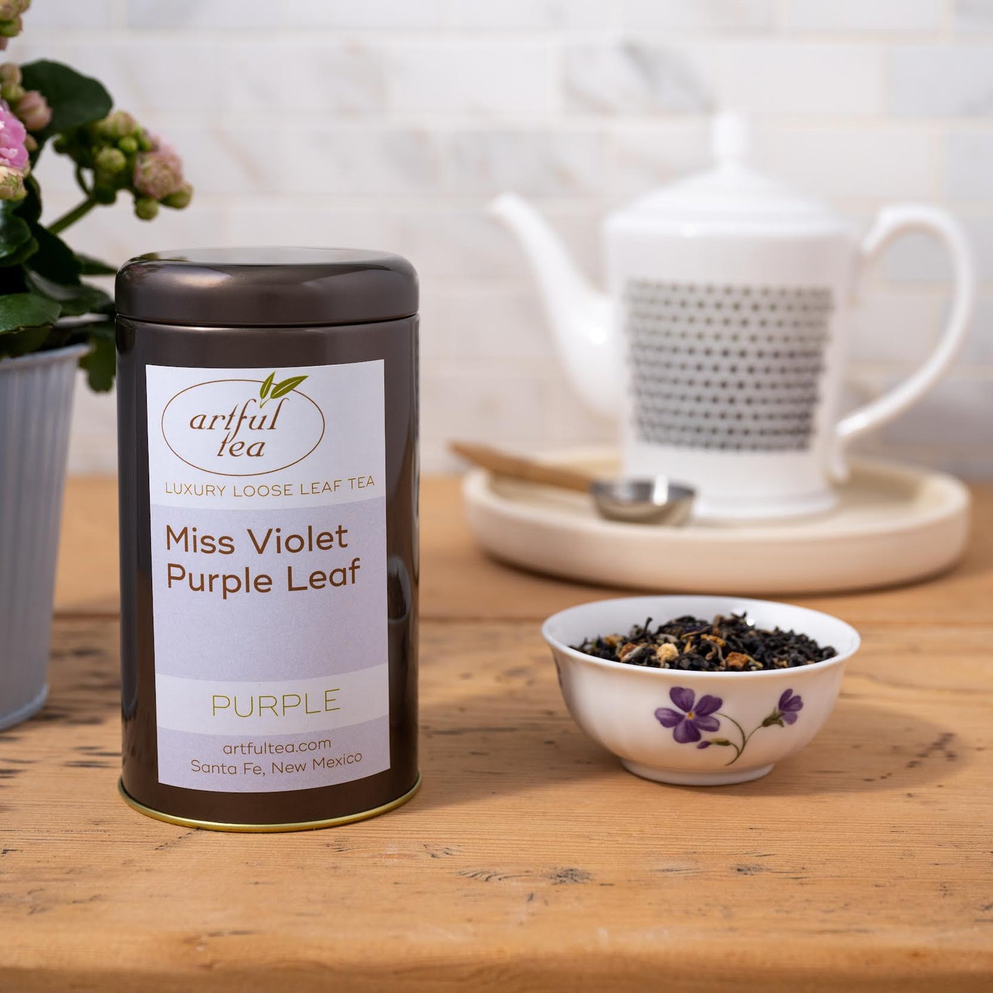 Miss Violet Purple Leaf Tea shown packaged in a brown tin next to a white bowl of loose tea leaves decorated with purple flowers. In the background is a white teapot and wooden handled table spoon on a white dish.