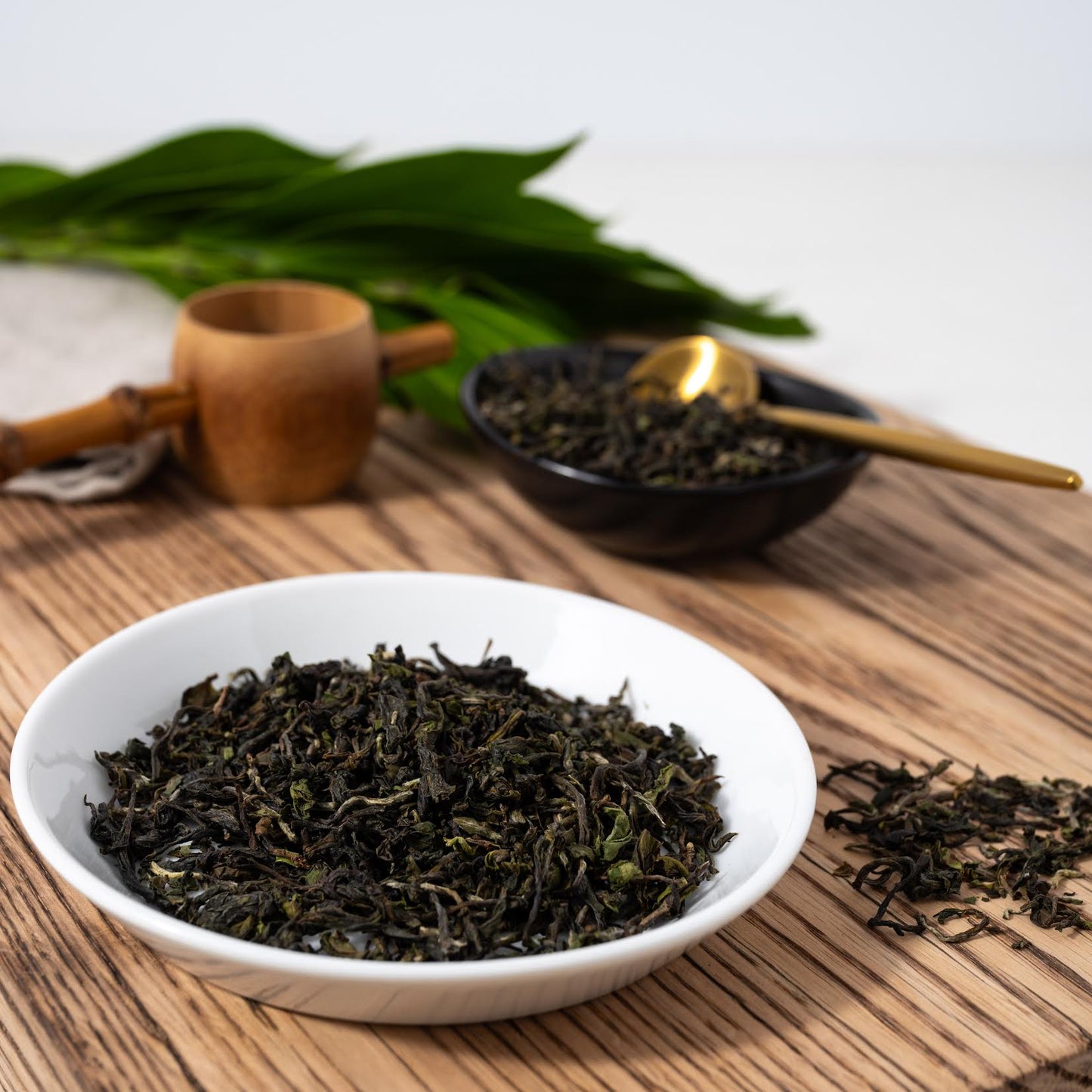 2025 First Flush Darjeeling loose tea leaves are shown in a white bowl as well as a black bowl with a gold spoon. A wooden scoop and loose tea leaves are nearby.