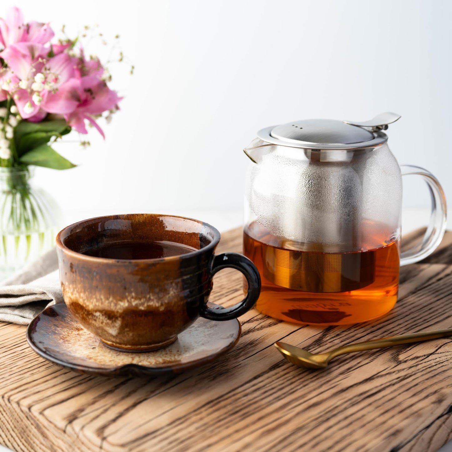 2025 First Flush Darjeeling Black Tea shown brewed in a clear teapot as well as in a brown and black mug on a matching dish. Flowers and a gold spoon are nearby.