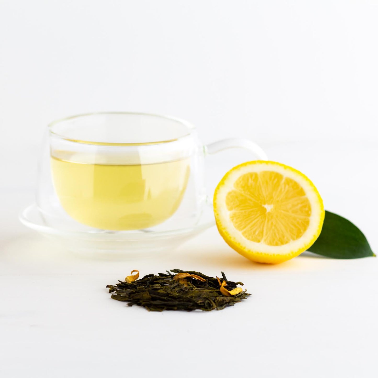 Green Tea Citrus shown brewed in a clear mug with loose tea leaves and a lemon nearby.
