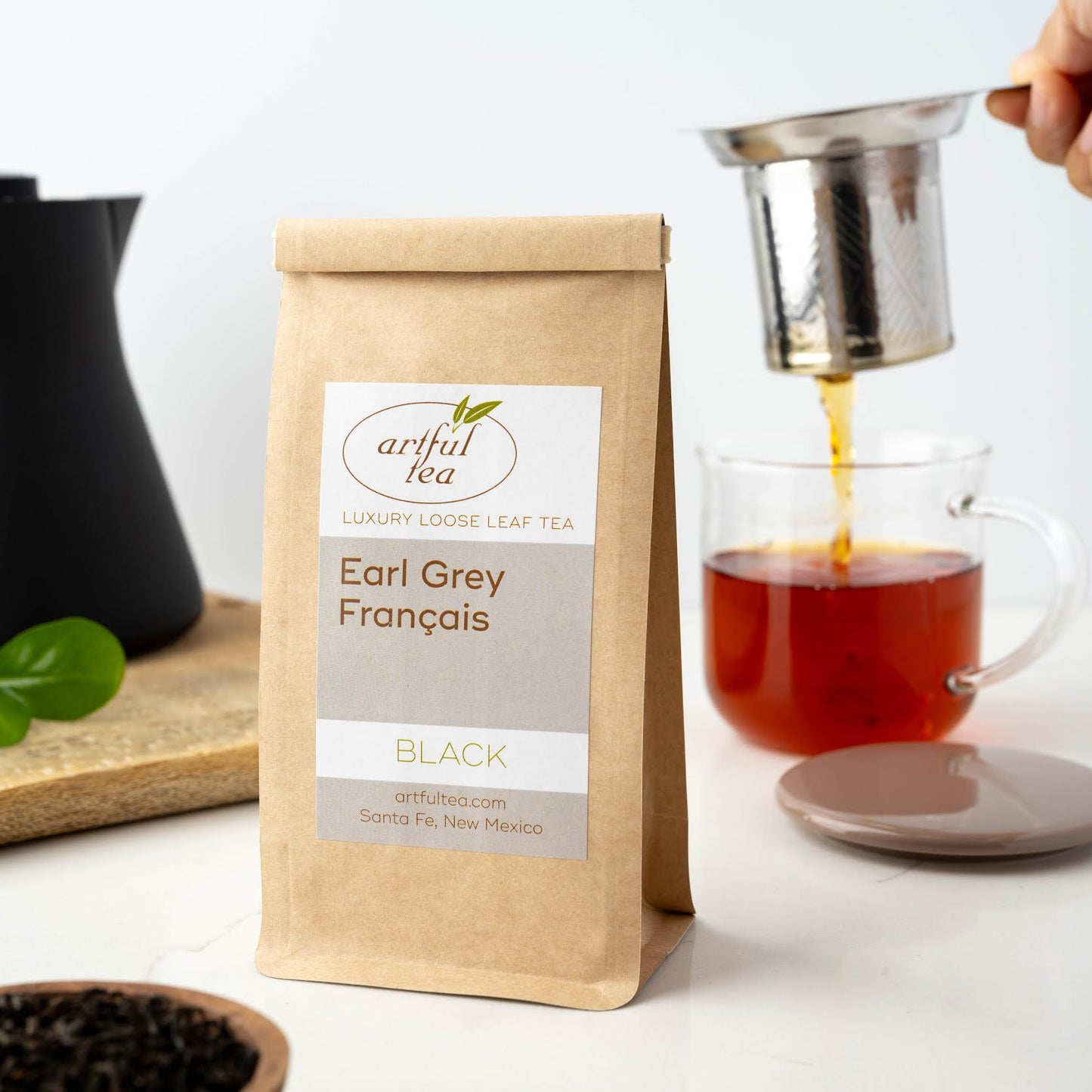 Earl Grey Français Black Tea shown packaged in a brown Kraft bag in front of a basket of tea leaves straining into a clear mug of brewed tea.