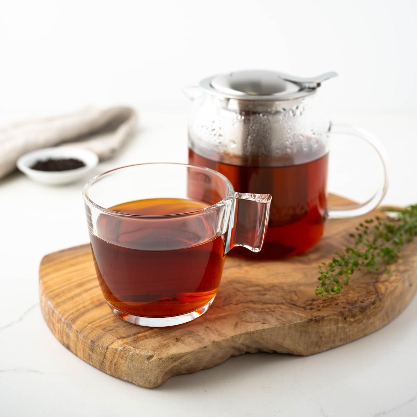 English Breakfast Black tea shown as brewed tea in a clear mug next to a clear teapot full of brewed tea, both on top of a wooden plank with ingredients nearby.