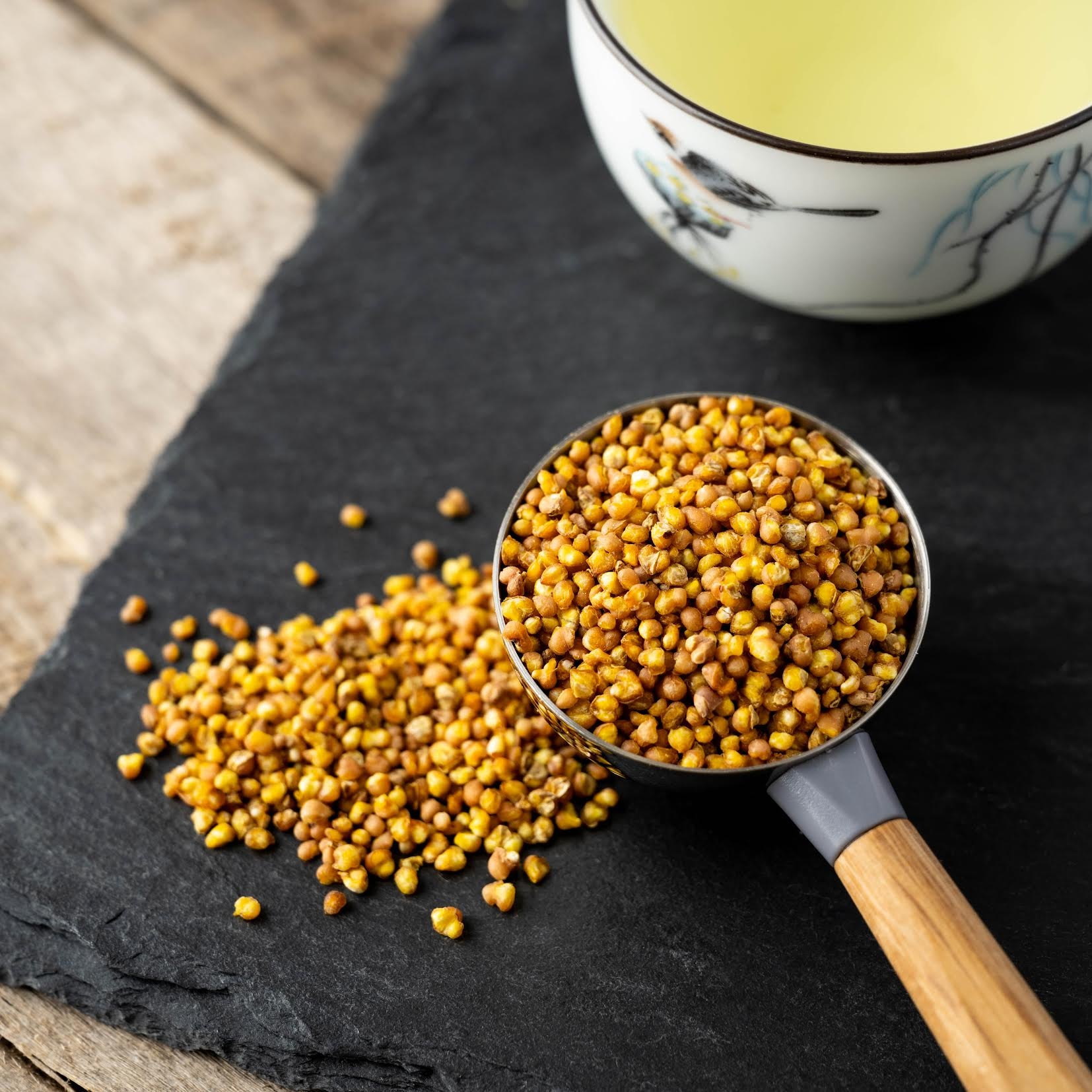Sobacha Buckwheat Tea shown as loose tea in teaspoon next to a pile of loose buckwheat tea on a black background.