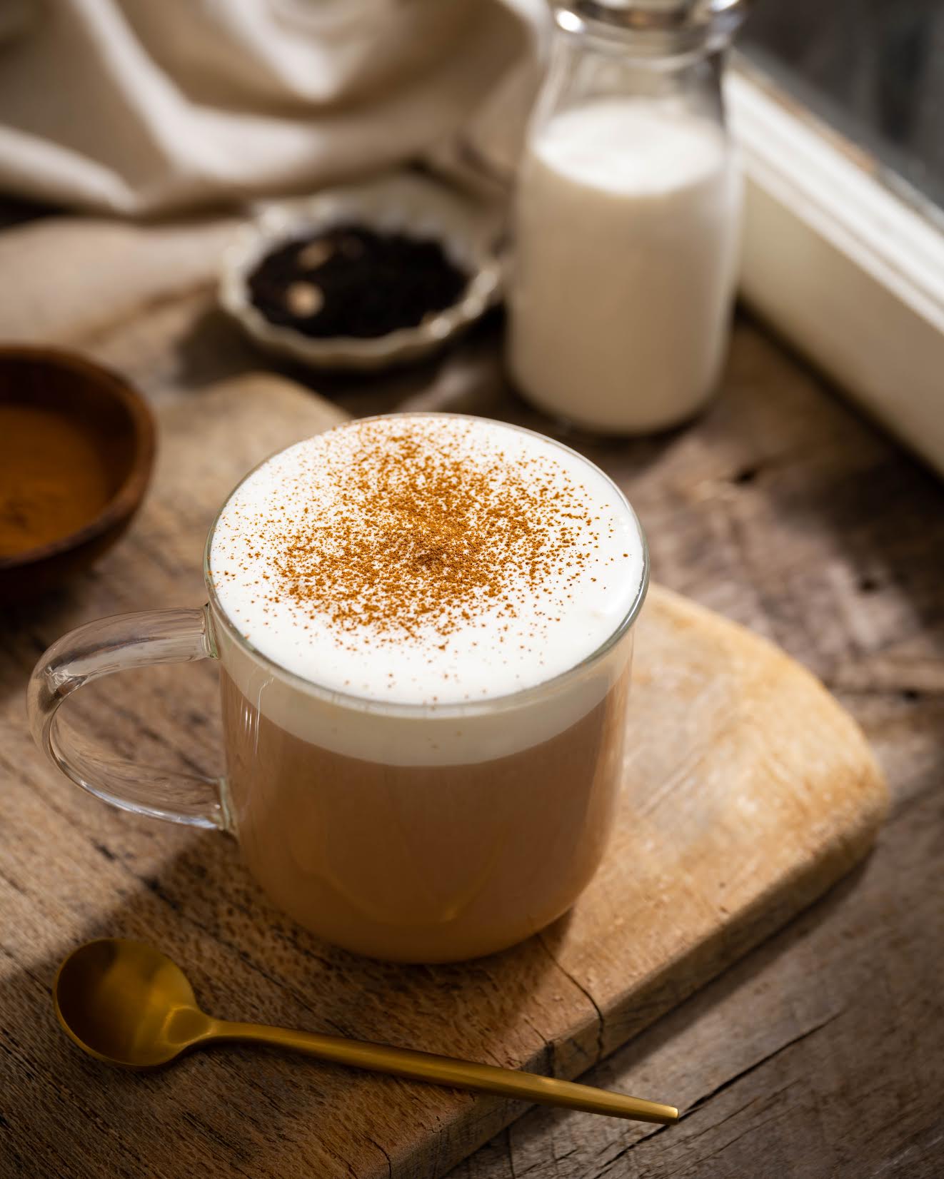 Caramel Pu-erh Tea shown brewed as a milky drink in a clear mug on wooden plank. Next to it is a golden spoon and in the background is a glass of milk and a dish of loose tea leaves.