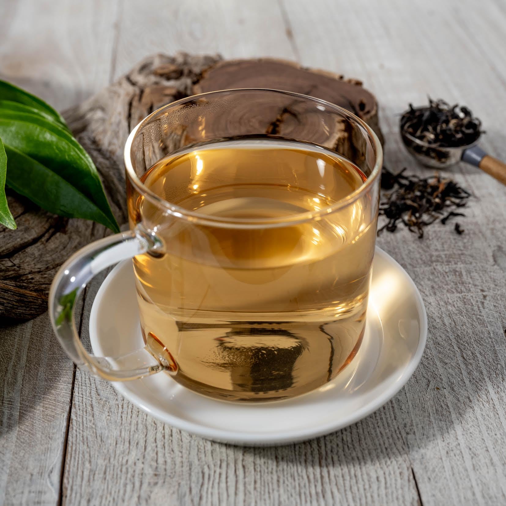 Kenyan Purple Leaf Tea shown as brewed tea in a clear mug on top of a white dish. In the background is a piece of wood and a tablespoon full of loose tea leaves.