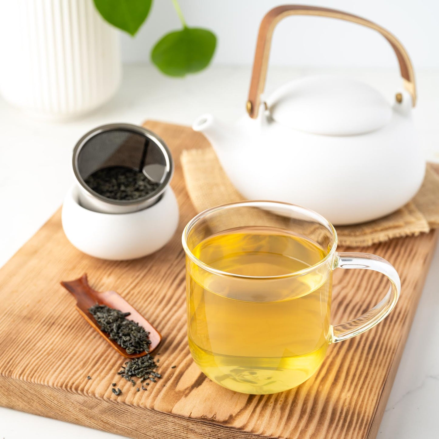 Chun Mee Green Tea shown brewed in a clear mug with loose tea leaves in a wooden scoop and strainer and a white teapot on burlap nearby.