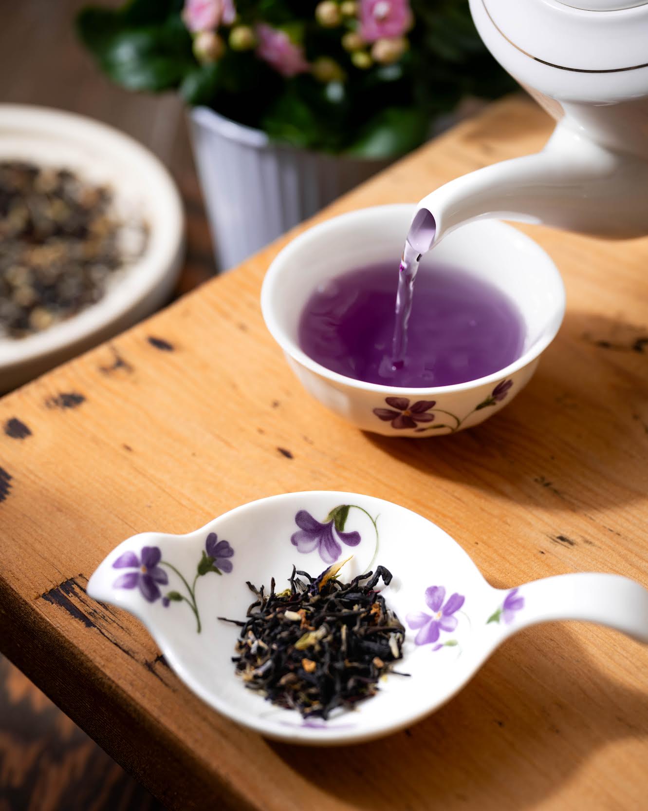 Miss Violet Purple Leaf Tea shown as brewed tea pouring out of a white teapot into a white cup with purple flowers. In front of it is a white dish full of loose tea leaves.