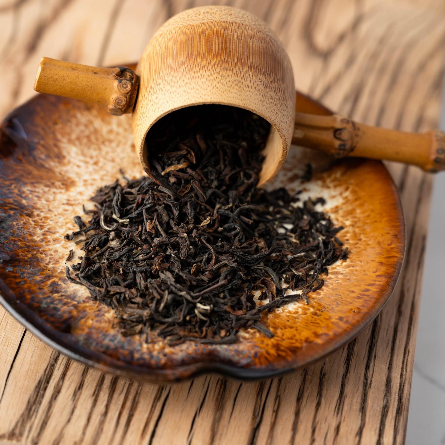 Daily Darjeeling loose black tea leaves spilling out of a wooden scoop onto a speckled brown plate on top of a wooden plank.