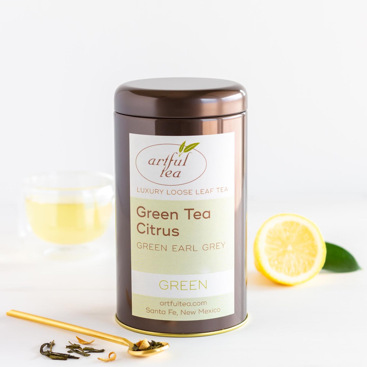 Green Tea Citrus shown packaged in a brown tin with a gold spoon of loose tea leaves in front and a clear mug of brewed tea and a lemon in the background.