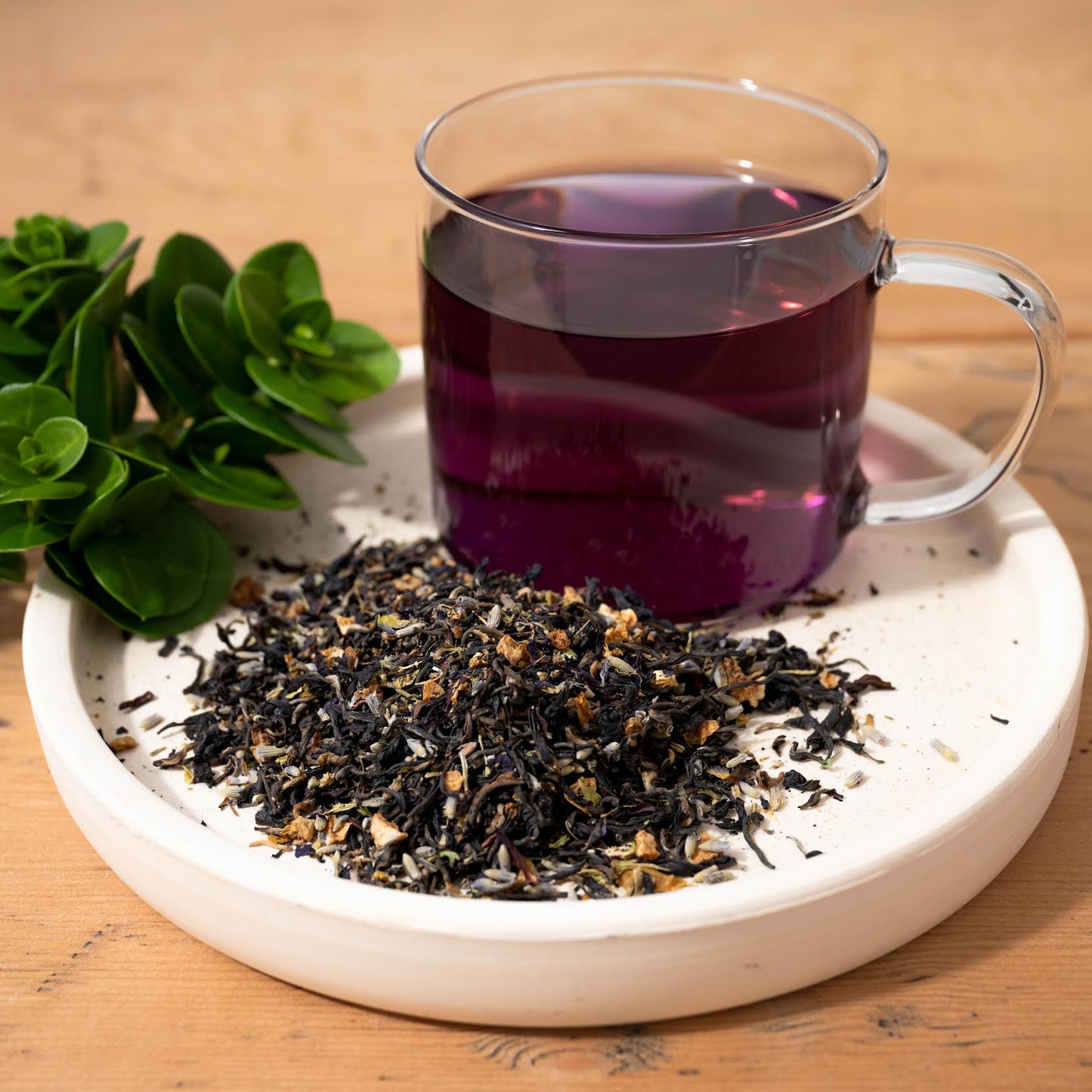 Miss Violet Purple Leaf Tea shown as brewed tea in a clear mug next to a pile of loose tea leaves, all on top of a white dish.