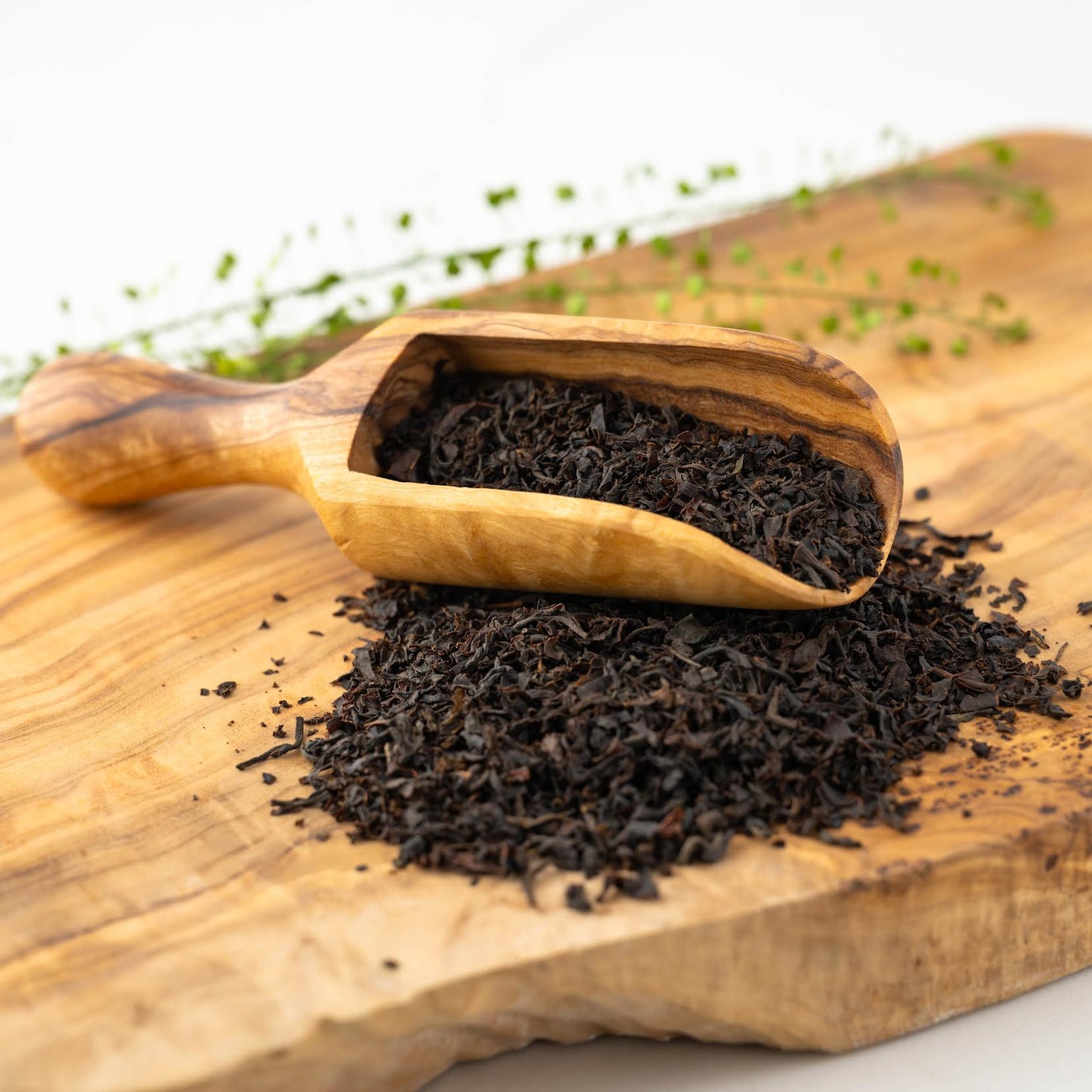 English Breakfast loose black tea leaves spilling out of a wooden scoop onto a wooden plank with ingredients nearby.