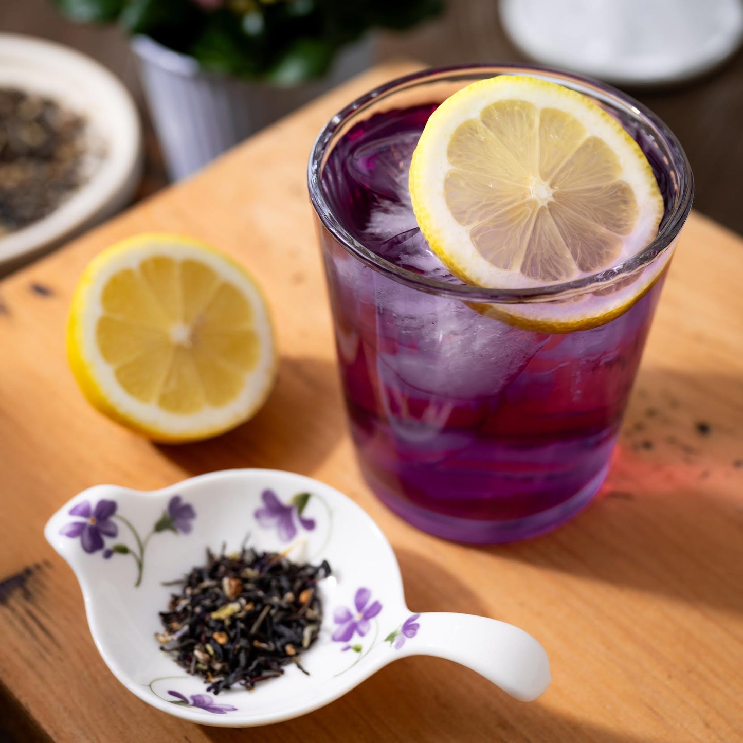 Miss Violet Purple Leaf Tea shown as brewed tea in a clear cup with slices of lemon. Next to it are loose tea leaves in a white dish decorated with purple flowers.