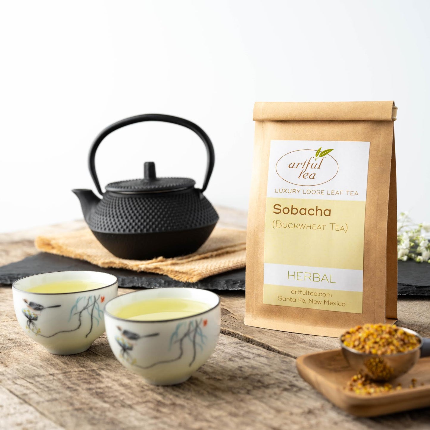 Sobacha Buckwheat tea shown packaged in a brown Kraft bag. In the foreground is a scoop of loose buckwheat tea next to two cups of brewed tea. In the background is a black teapot on burlap.