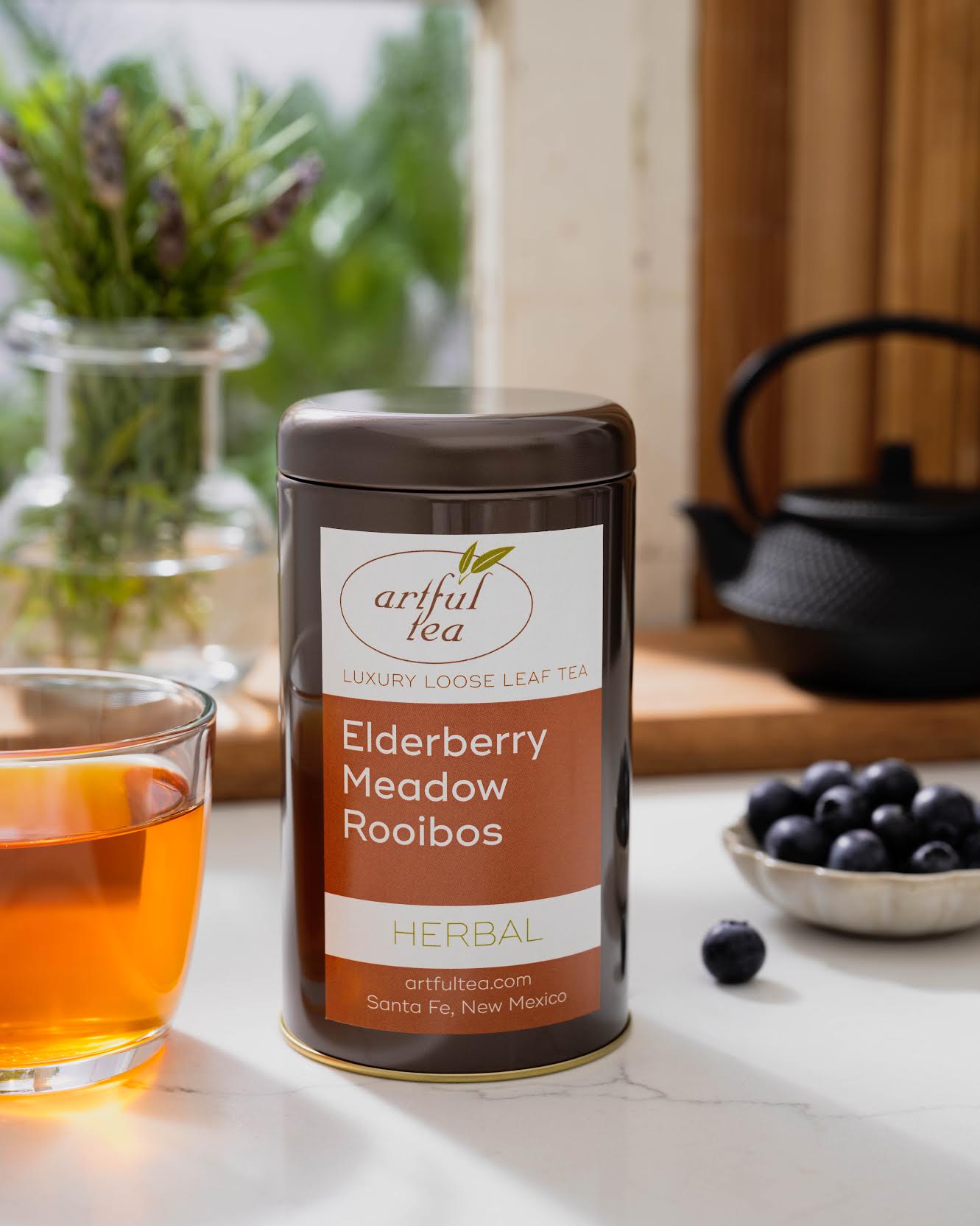 Elderberry Meadow Rooibos Herbal Tea shown packaged in a brown tin next to a cup of brewed tea and a small bowl of blueberries. In the background is a clear vase of flowers and a black teapot in a window sill.