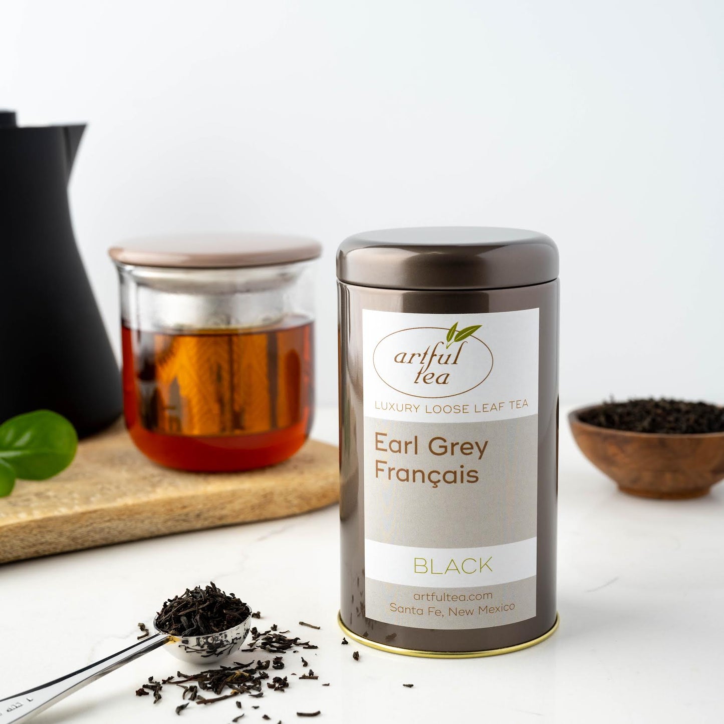 Earl Grey Français shown packaged in a brown tin next to a silver scoop of loose leaf tea. In the background is a clear mug full of brewed tea and a dish full of tea leaves.