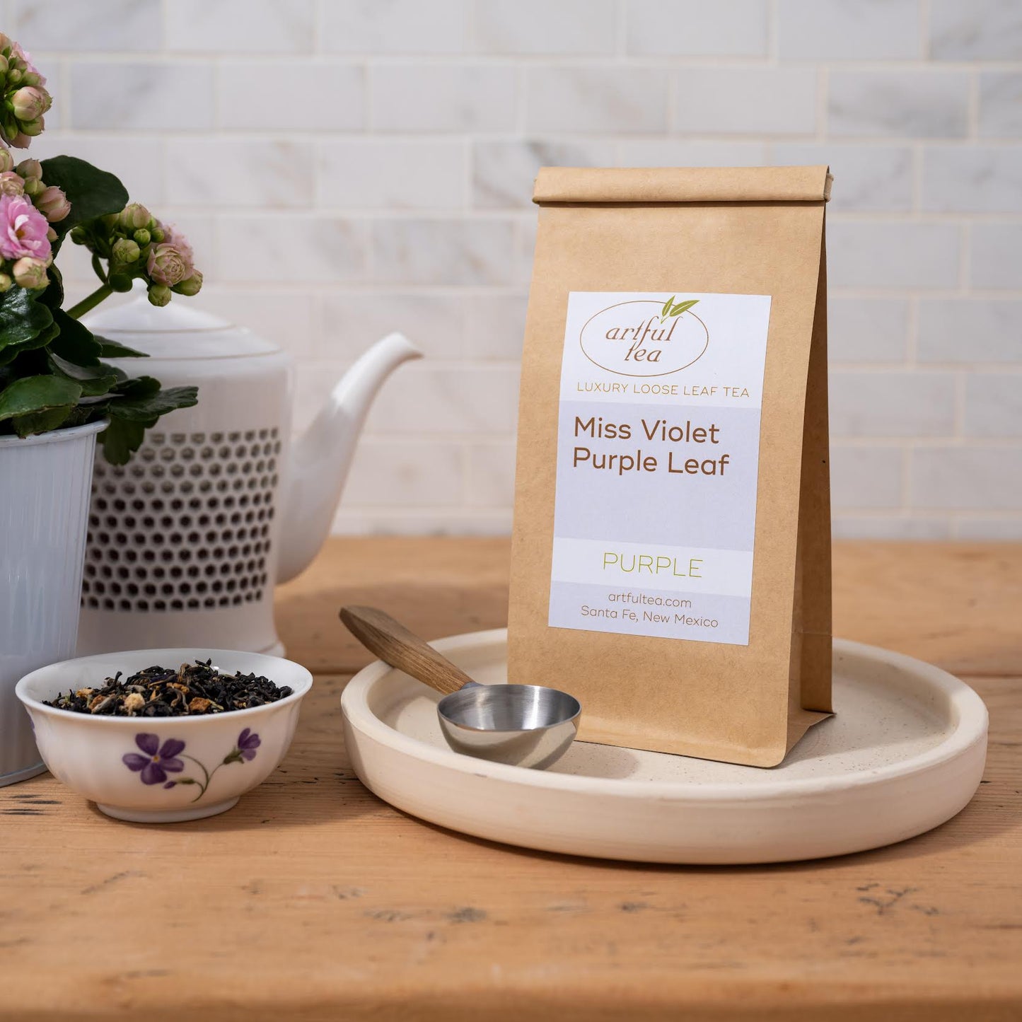 Miss Violet Purple Leaf Tea shown packaged in a brown Kraft bag and a table spoon on a white dish. Next to it is a white bowl full of loose tea leaves and a white tea pot.