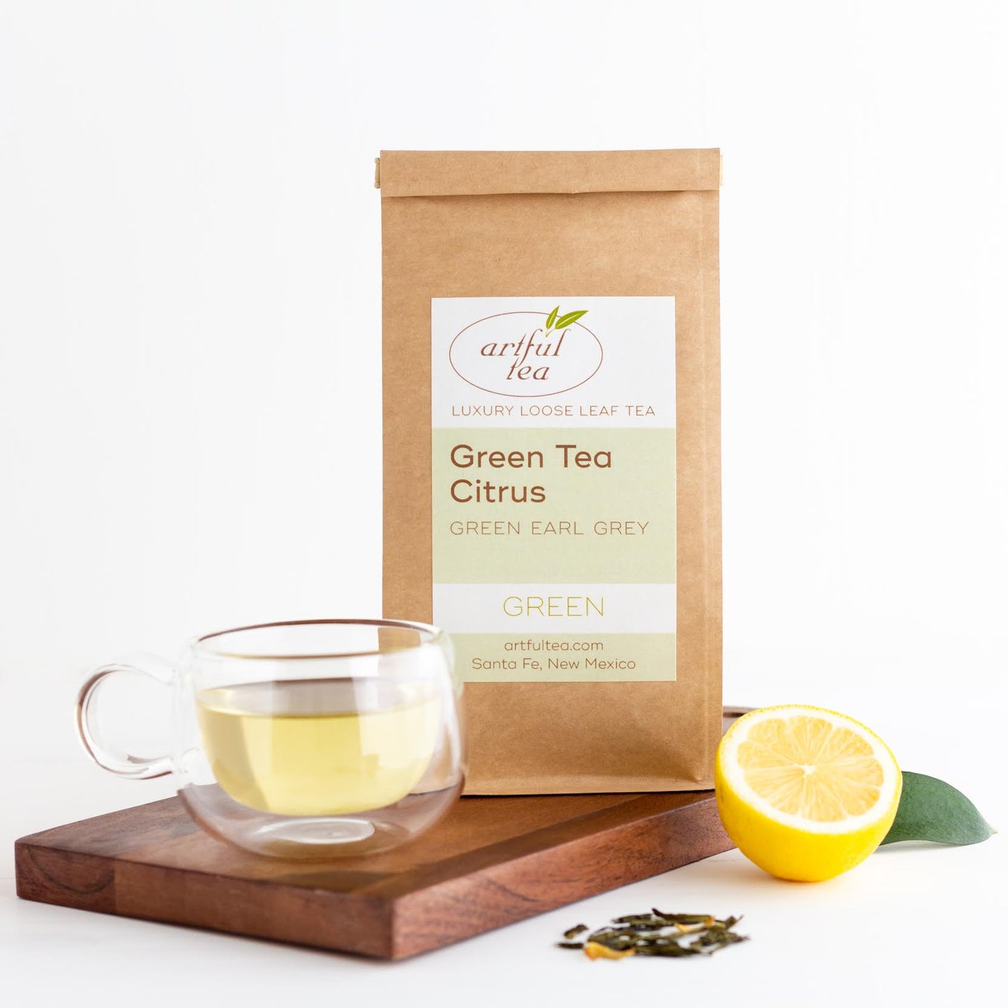 Green Tea Citrus shown packaged in a brown Kraft bag behind a clear mug of brewed tea on a wooden plank with loose tea leaves and a lemon nearby.