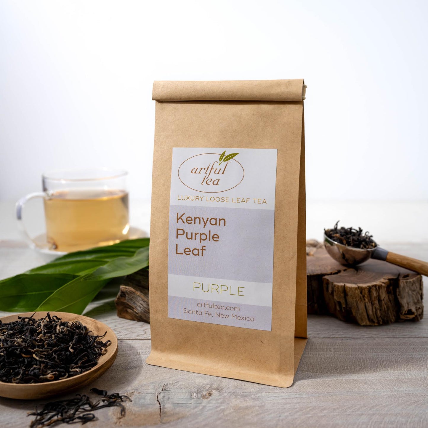 Kenyan Purple Leaf Tea shown packaged in a brown Kraft bag next to a wooden dish of loose tea leaves. In the background is a wooden handled tablespoon full of tea leaves on top of a piece of wood, as well as a clear mug full of brewed tea.