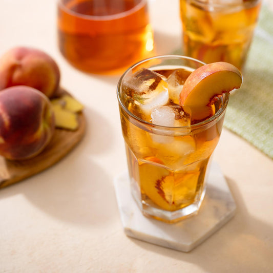Ginger Peach Black Tea shown as iced tea with a slice of a peach inside a clear cup on top of a marble coaster. Other clear cups full of tea and peaches nearby.