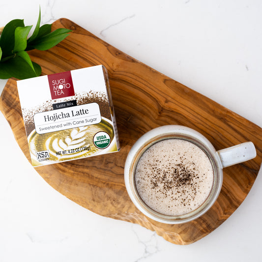 Hojicha Latte shown packaged in a box next to a mug of hot Hojicha Latte on a wooden plank.