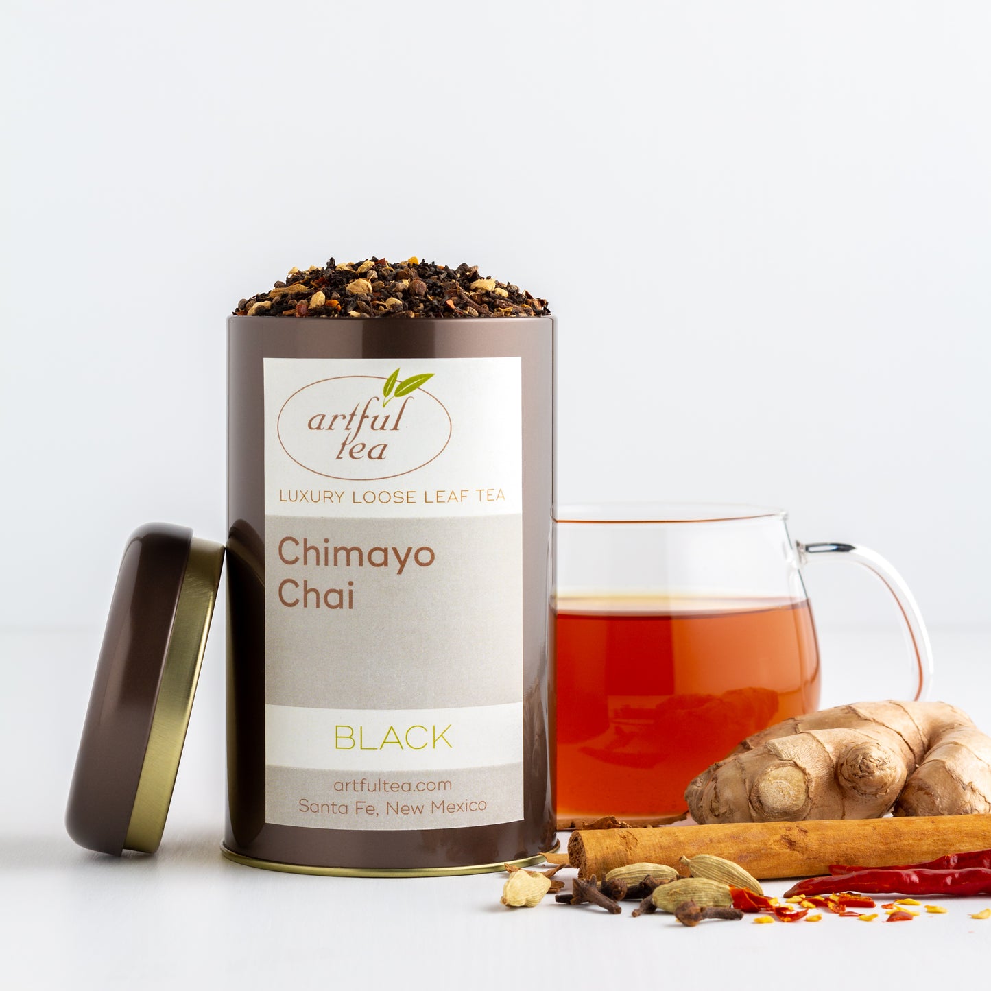 Chimayo Chai Black Tea shown packaged in a brown tin with the lid off. A glass mug of brewed tea is in the background, with ginger root, cinnamon sticks, cardamom pods, and chile peppers nearby.