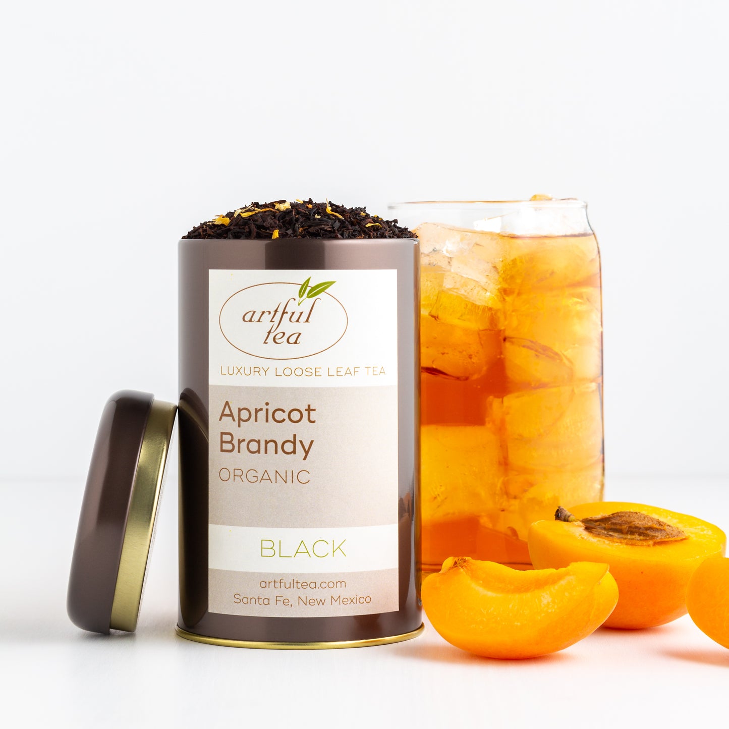 Apricot Brandy Organic Black Tea shown packaged in a brown tin with the lid off, next to a tall glass of iced tea and a freshly sliced apricot