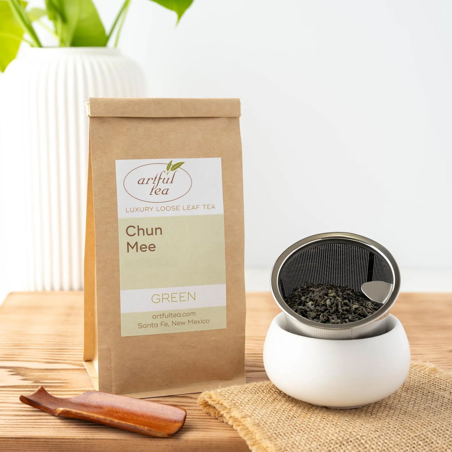 Chun Mee Green Tea shown packaged in a brown Kraft bag next to a wooden scoop and loose tea leaves in a strainer on burlap.