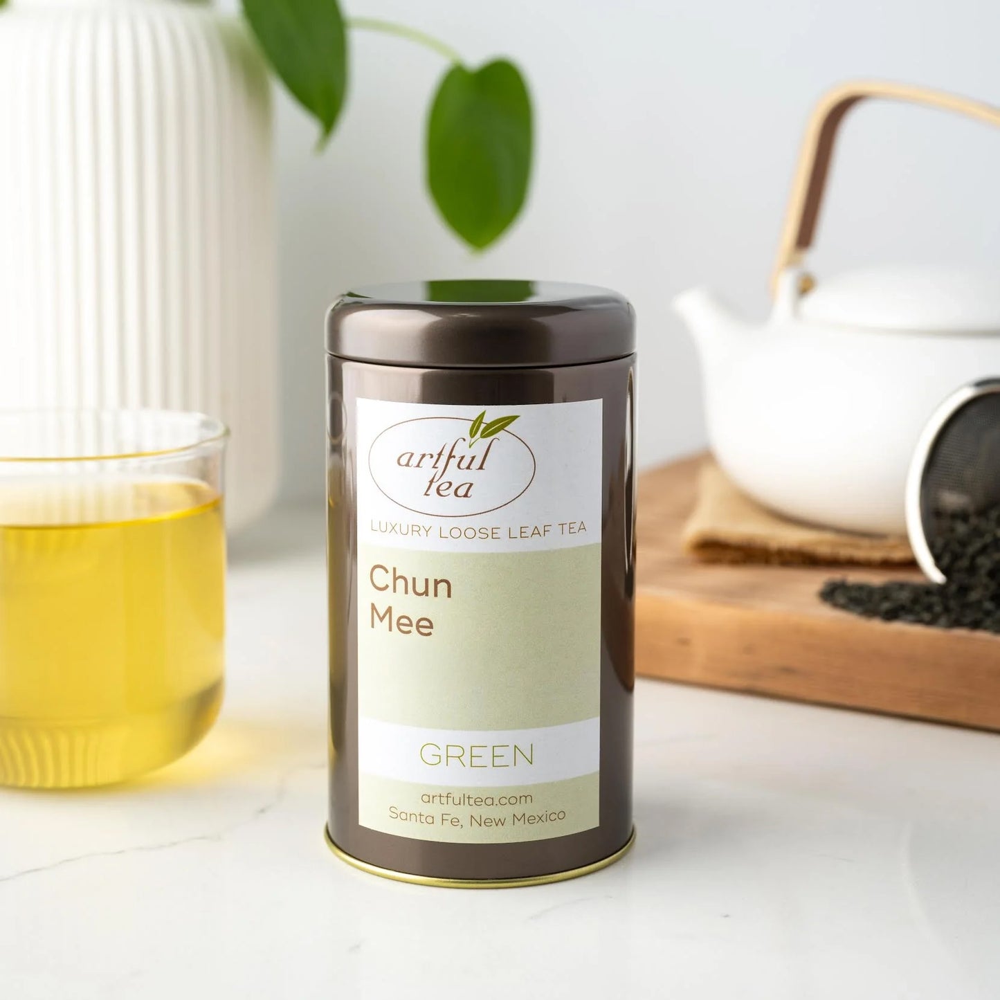 Chun Mee Green Tea shown packaged in a brown tin next to a clear cup of brewed tea. In the background is a white teapot and a strainer with tea leaves spilling out on top of a wooden plank.