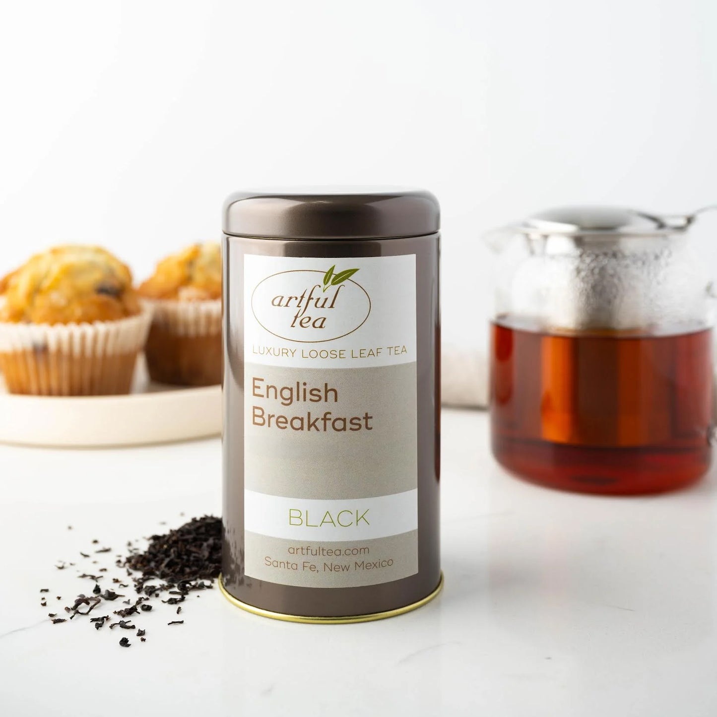 English Breakfast Black Tea shown packaged in a brown tin with loose tea leaves next to it. In the background is a clear teapot full of brewed tea and muffins on a white tray.