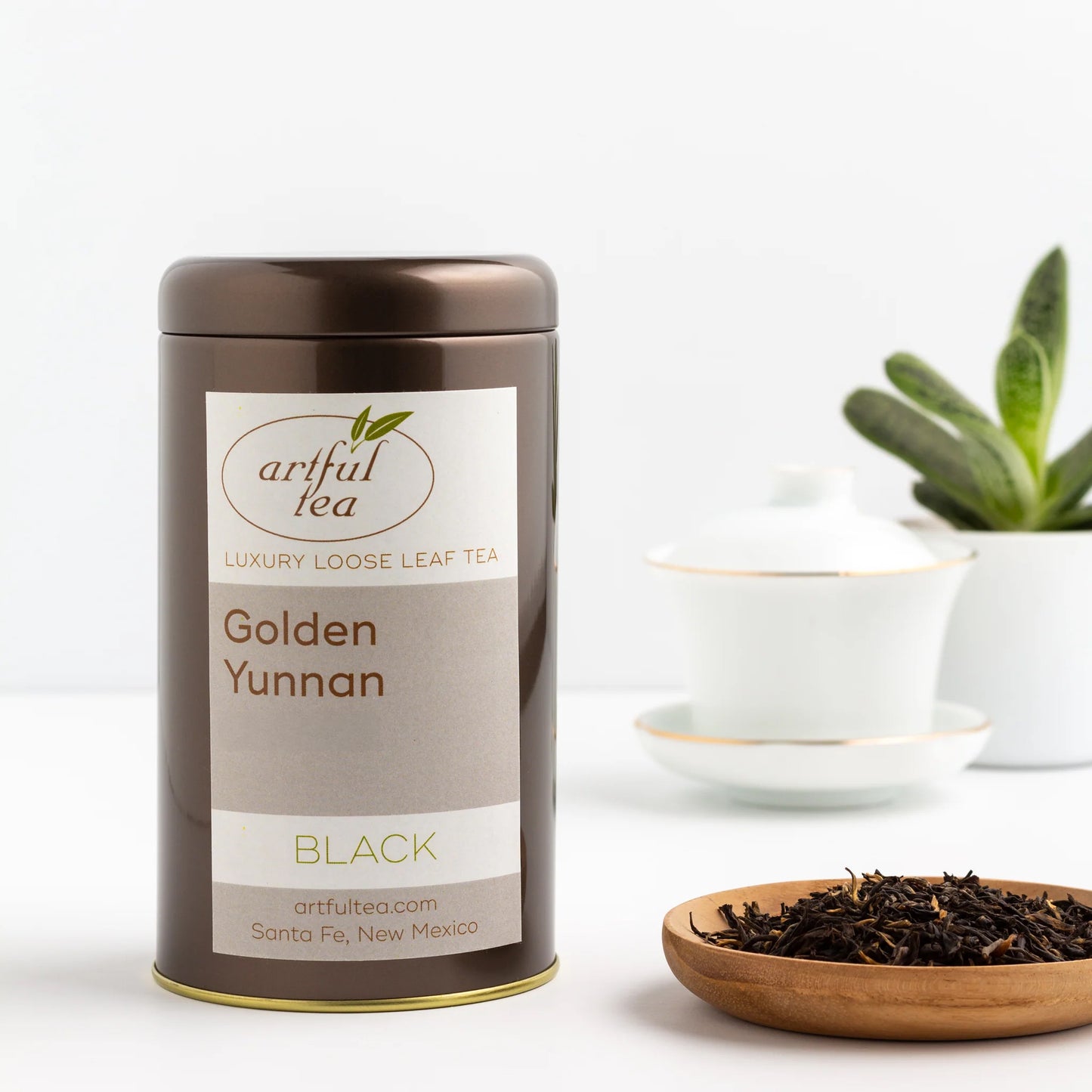 Golden Yunnan Black Tea show in a brown tin next to a wooden bowl full of loose black tea leaves. In the background is a white teacup with a gold rim on a matching plate and a plant in a white pot.