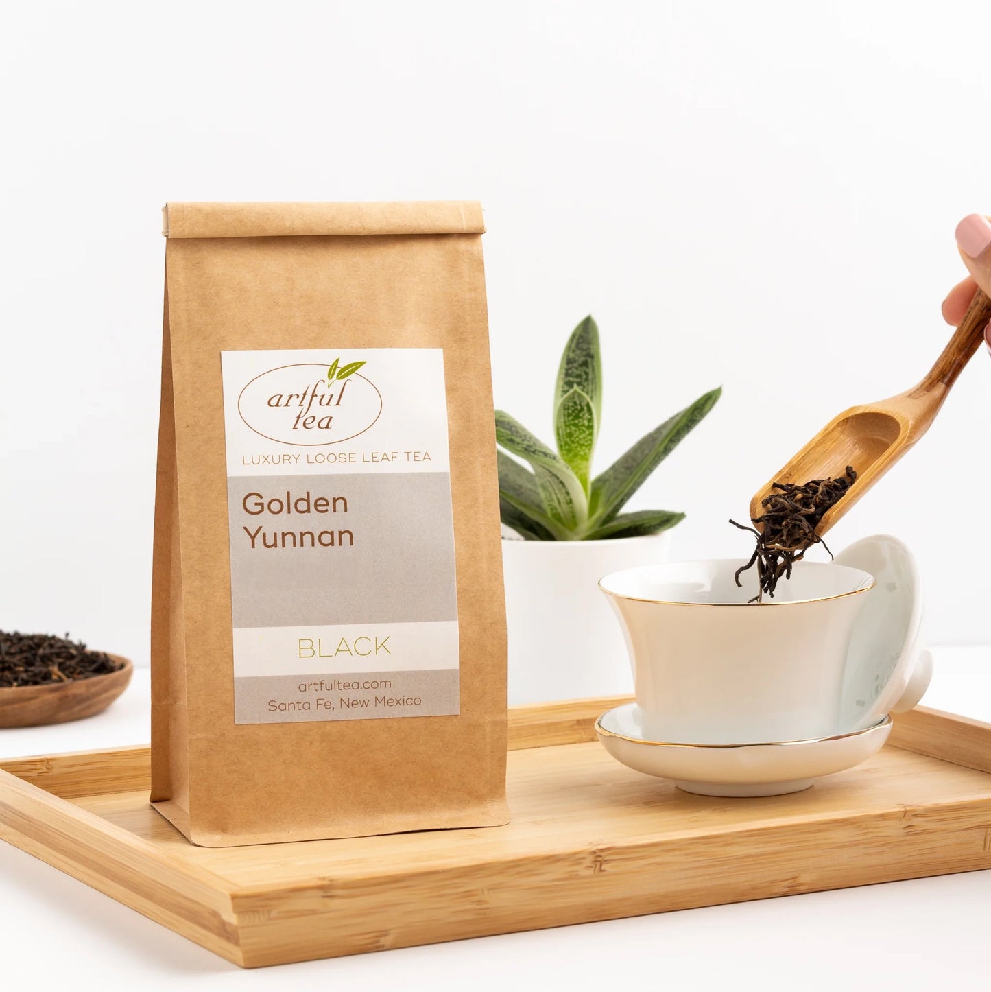 Golden Yunnan Black Tea shown in a brown Kraft bag next to a white teacup with a gold rim on a matching plate with a wooden scoop of loose tea leaves being poured into it, all on top of a wooden tray.