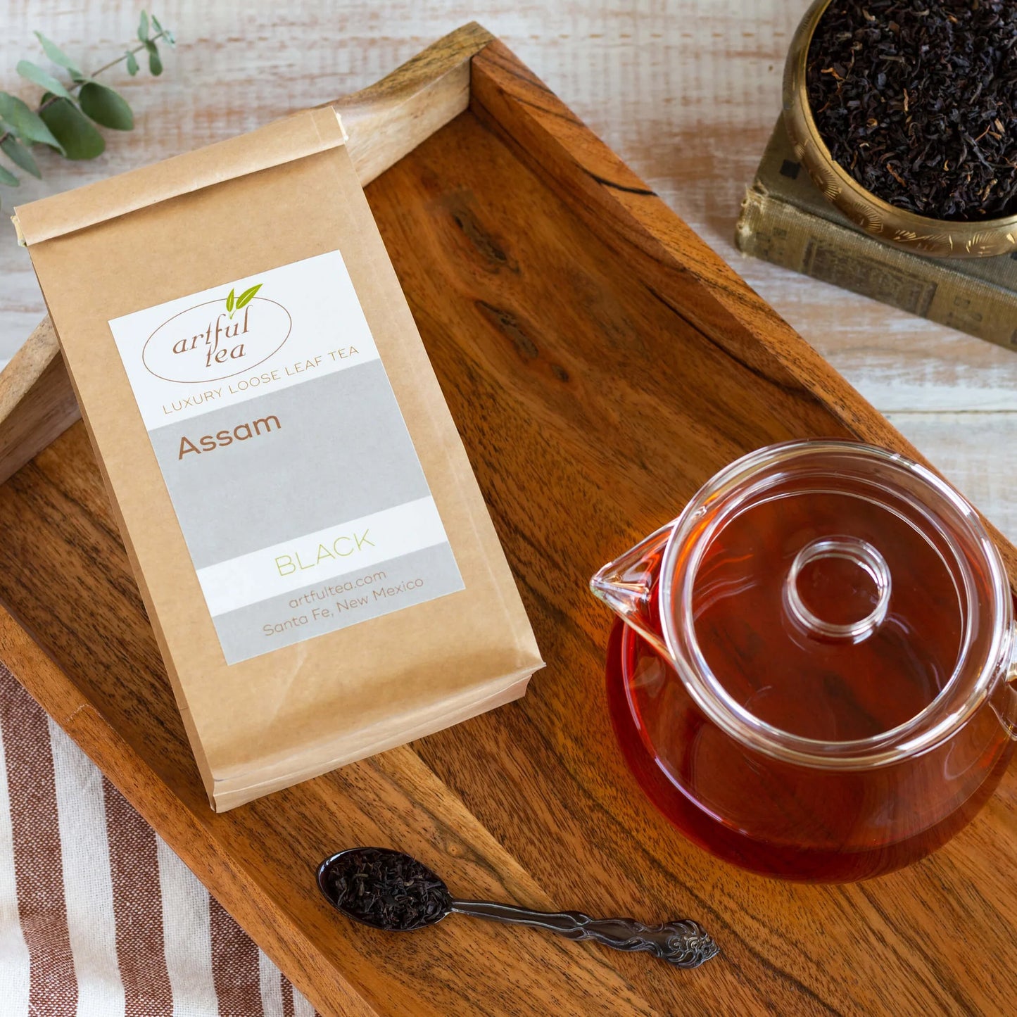 Assam Black Tea shown packaged in a brown Kraft bag next to a spoon of tea leaves and a clear teapot full of brewed tea all on top of a wooden tray.