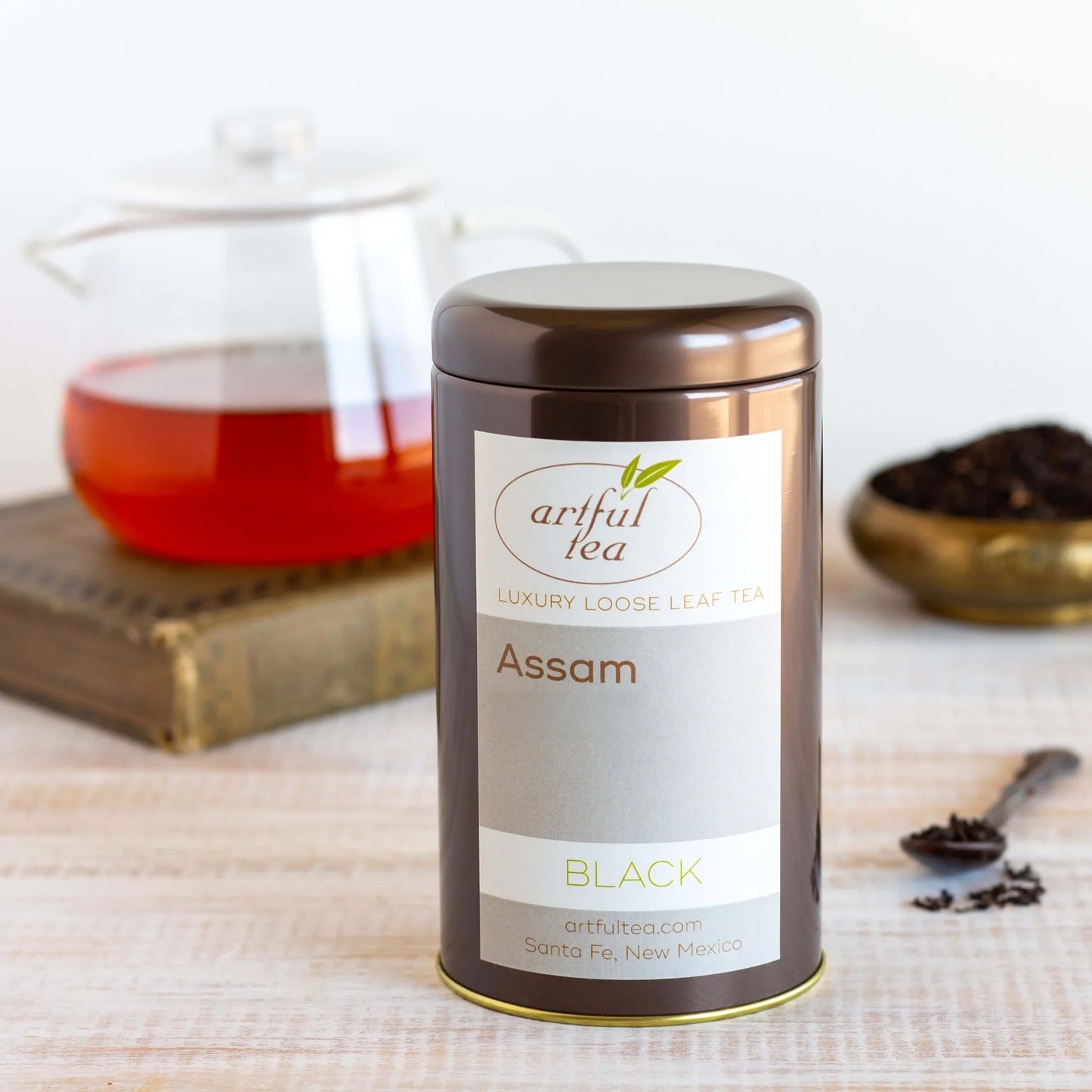 Assam Black Tea shown packaged in a brown tin in front of a clear teapot full of brewed tea on top of a book as well as a brass dish and spoon full of loose tea leaves.