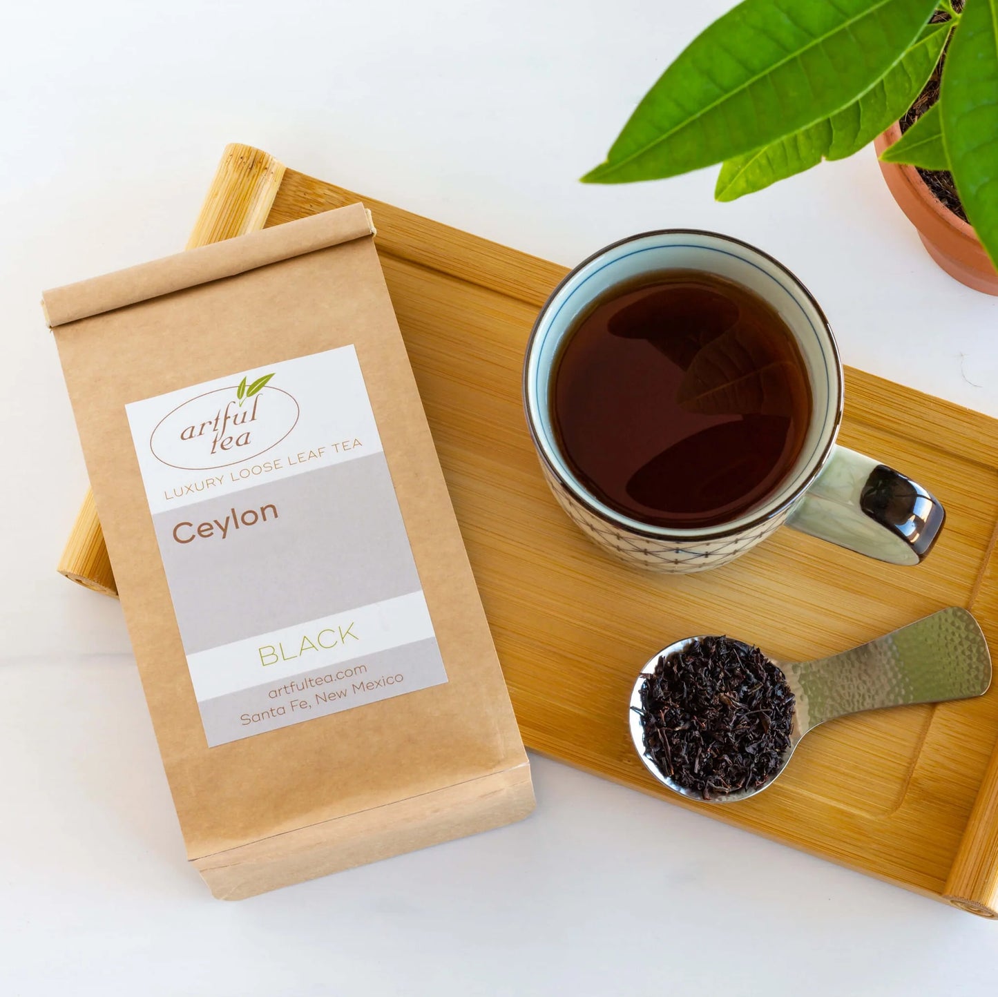 Ceylon Black Tea shown packaged in a brown Kraft bag next to a mug full of brewed tea and a silver scoop of loose tea leaves all on top of a wooden tray.