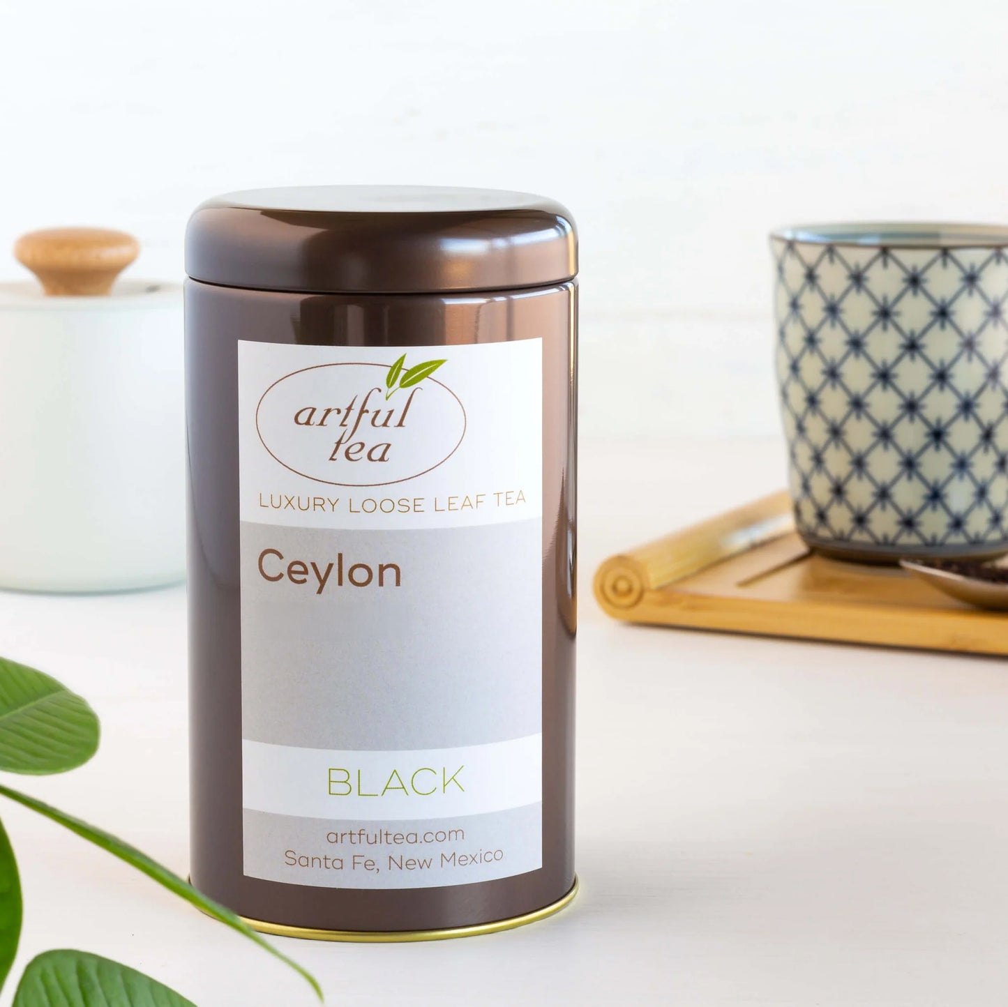 Ceylon Black Tea shown packaged in a brown tin in front of a white dish and a decorated cup on top of a wooden tray.