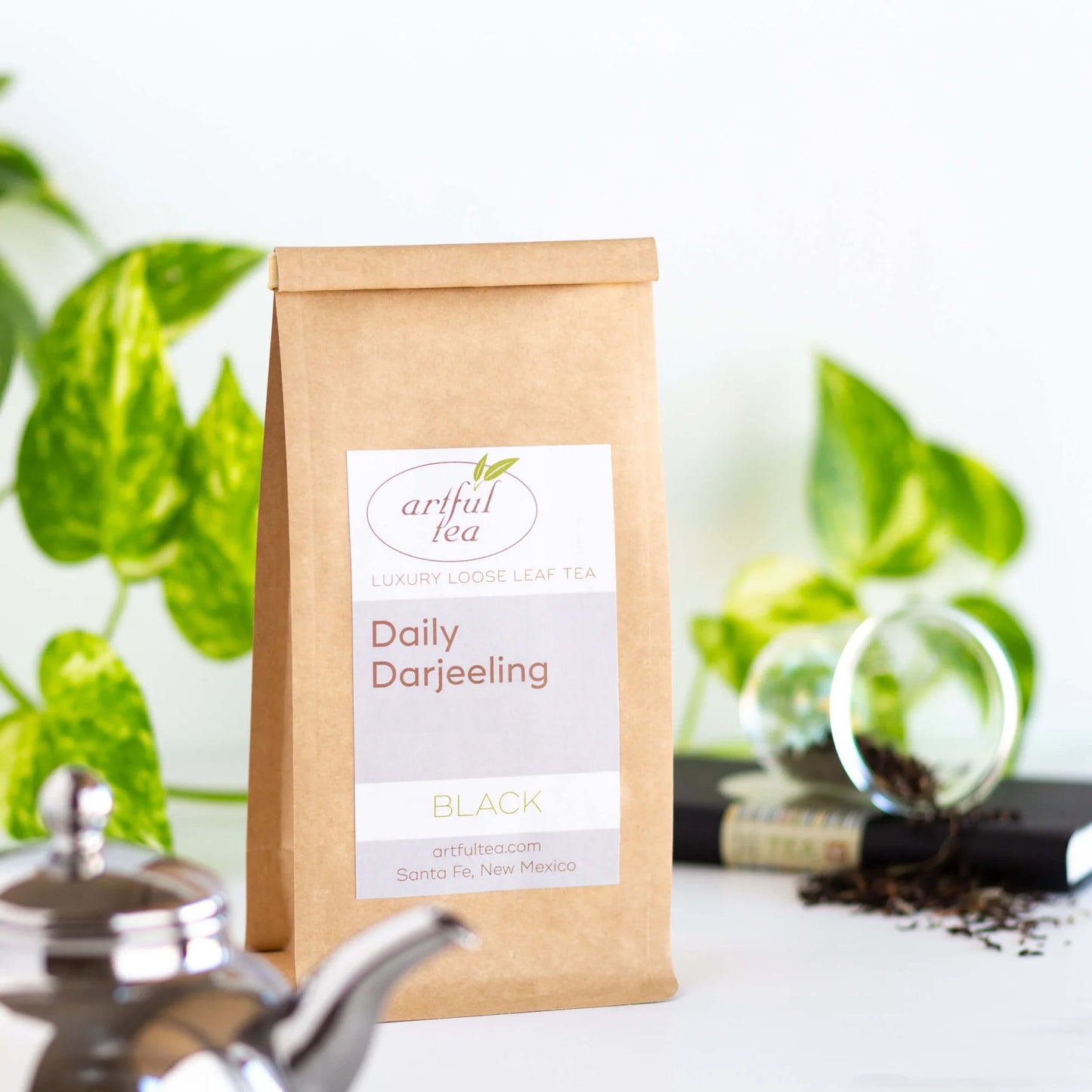 Daily Darjeeling Black Tea shown packaged in brown Kraft bag. Surrounding it is a clear cup of loose tea leaves spilling out on to a book, a silver teapot, and green plant leaves.