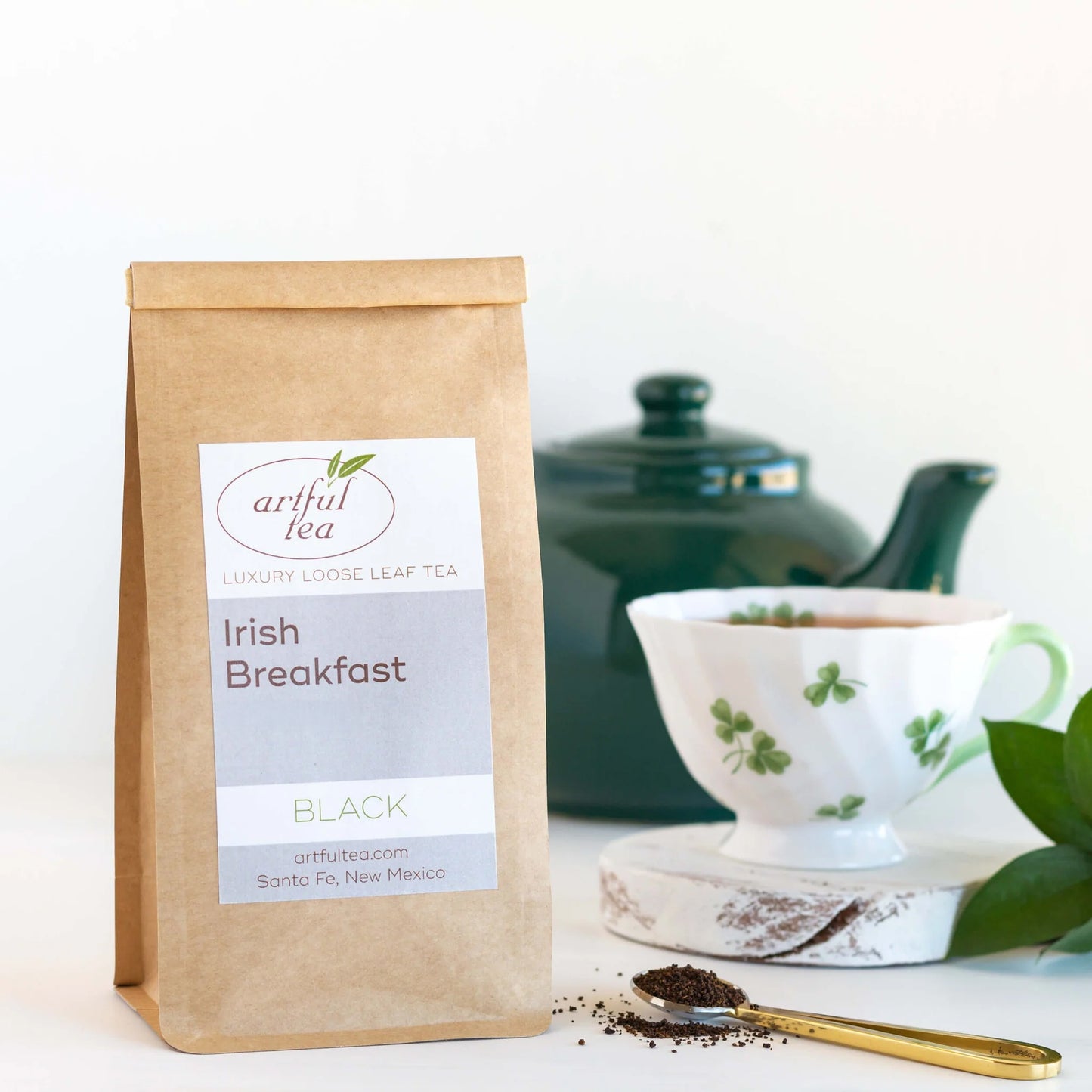 Irish Breakfast Black Tea shown in a brown Kraft bag next to a golden spoon full of loose black tea leaves. In the background is a white mug with green clovers full of brewed tea, and a dark green teapot.
