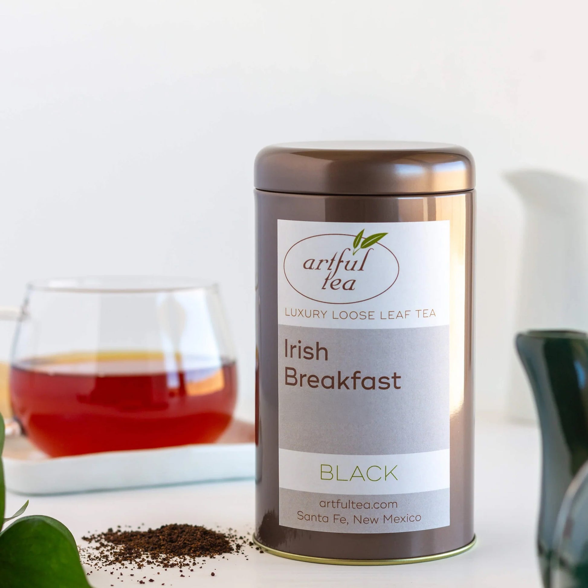 Irish Breakfast Black Tea shown in a brown tin with loose tea leaves next to it. In the background is a clear cup full of brewed tea.