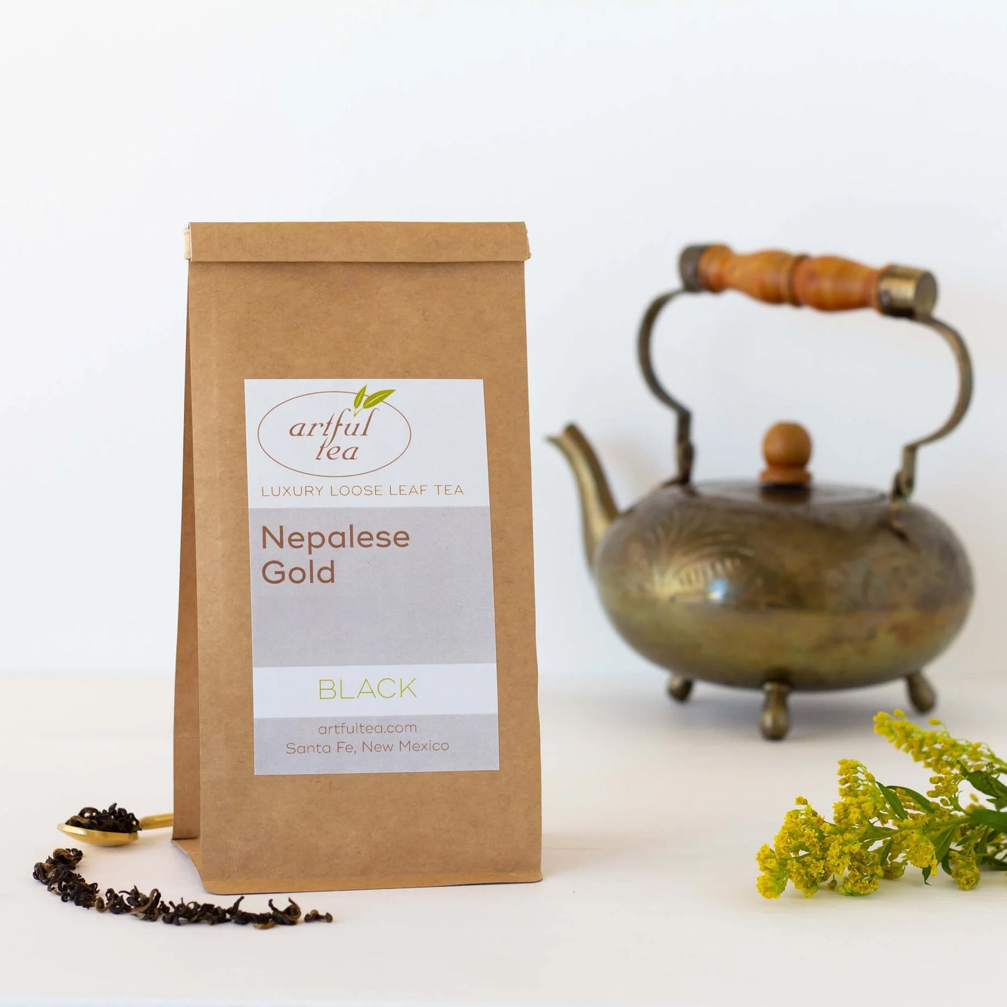 Nepalese Gold Black Tea show in a brown Kraft bag. Beside it is a gold spoon of loose tea leaves with the leaves spilling out and yellow flowers. In the background is a gold tea pot with a wooden handle.