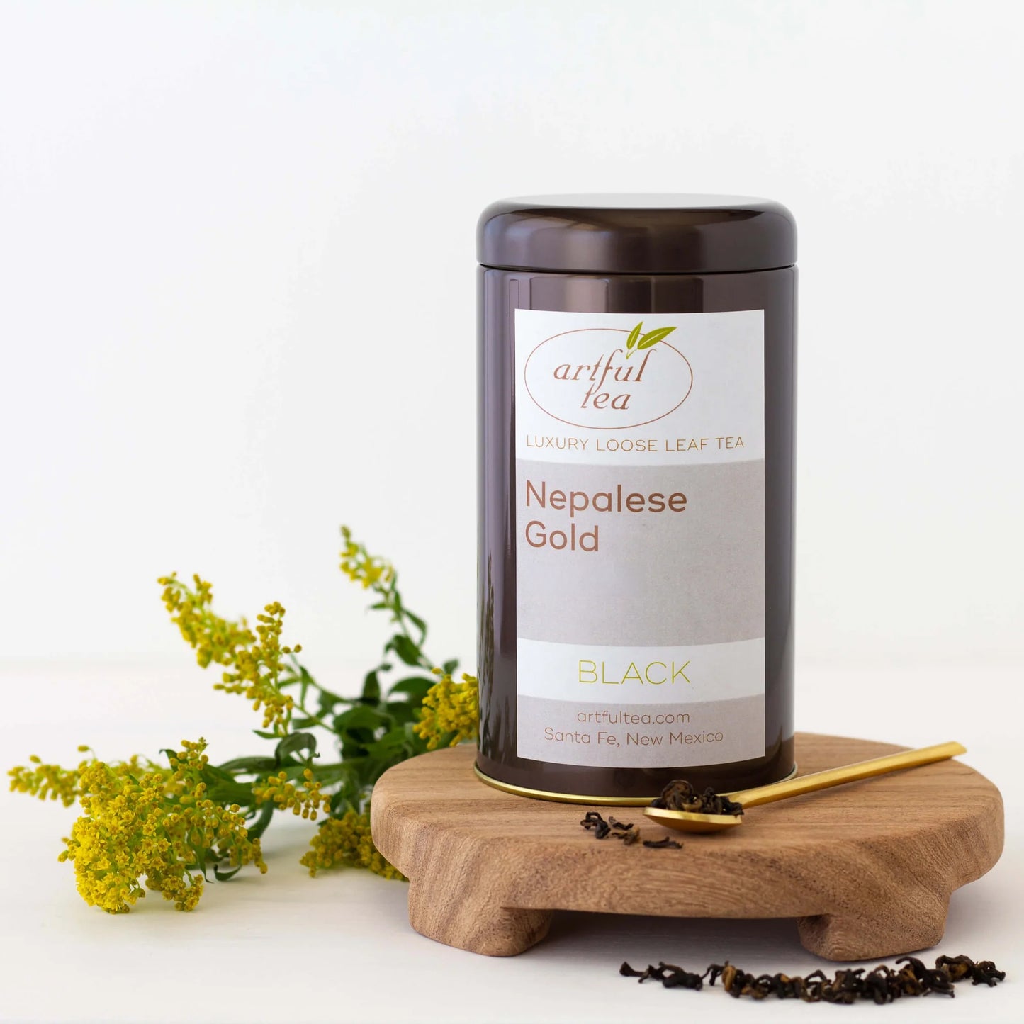 Nepalese Gold Black Tea show in a brown tin on a wooden coaster beside a gold spoon full of loose tea leaves. Next to it are more loose tea leaves and yellow flowers.