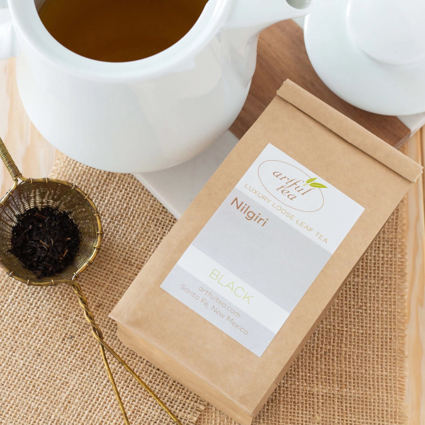 Nilgiri Black Tea shown packaged in a brown Kraft bag on burlap squares. Surrounding it is a golden basket of tea leaves and a white teapot full of brewed tea.