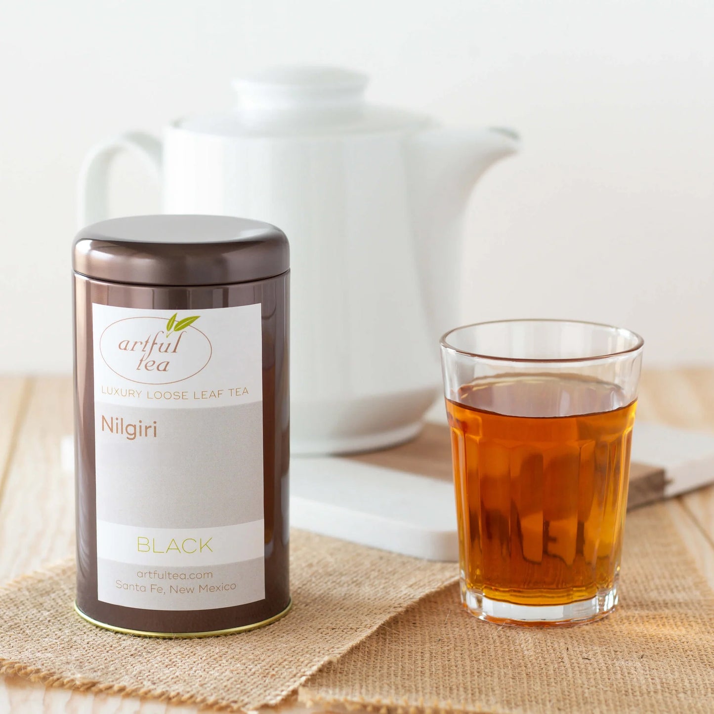 Nilgiri Black Tea shown packaged in a brown tin next to a cup of brewed tea on top of squares of burlap. In the background is a tall white teapot.