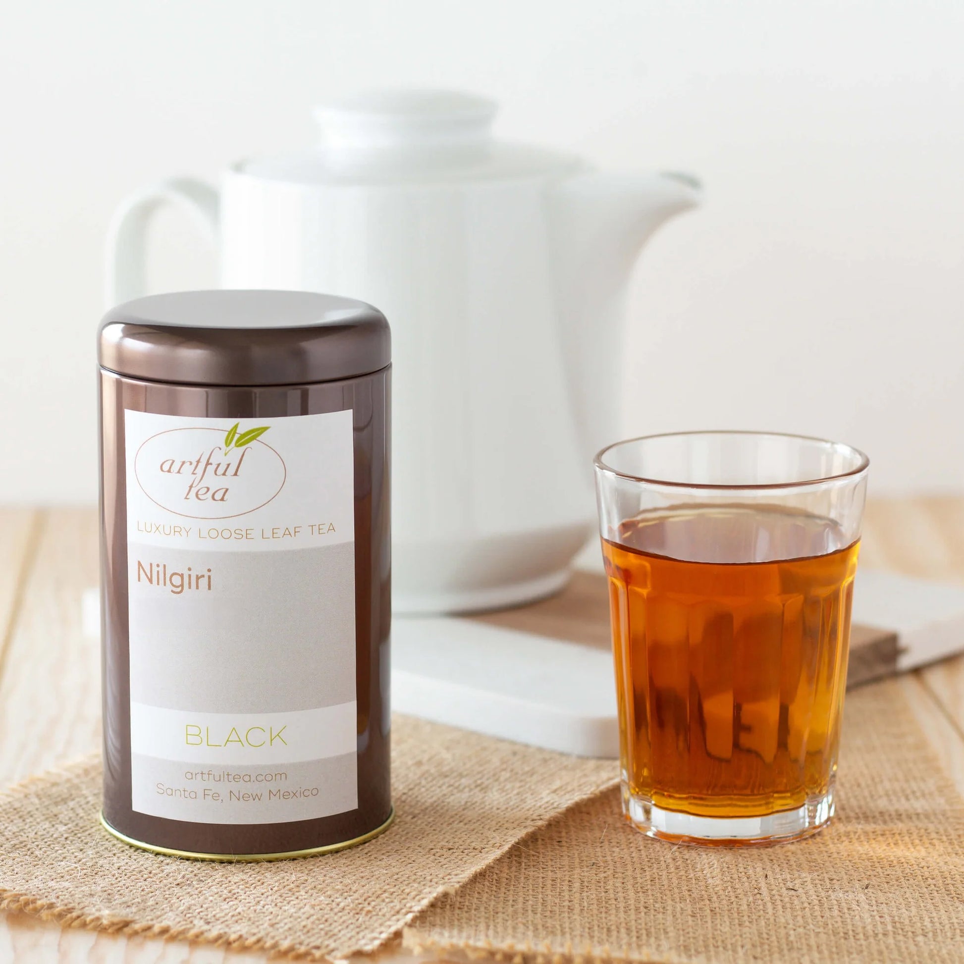Nilgiri Black Tea shown packaged in a brown tin next to a cup of brewed tea on top of squares of burlap. In the background is a tall white teapot.
