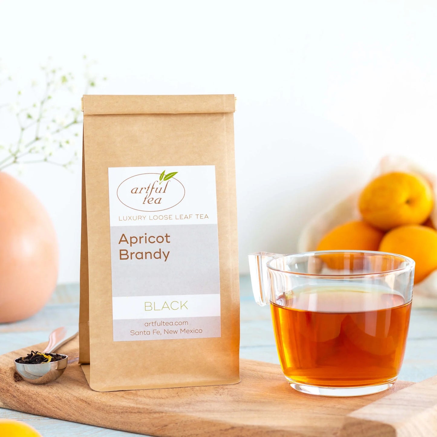 Apricot Brandy Black Tea shown packaged in a brown Kraft bag next to a clear mug of brewed tea and a silver teaspoon of loose leaf tea all on top of a wooden tray with ingredients nearby.