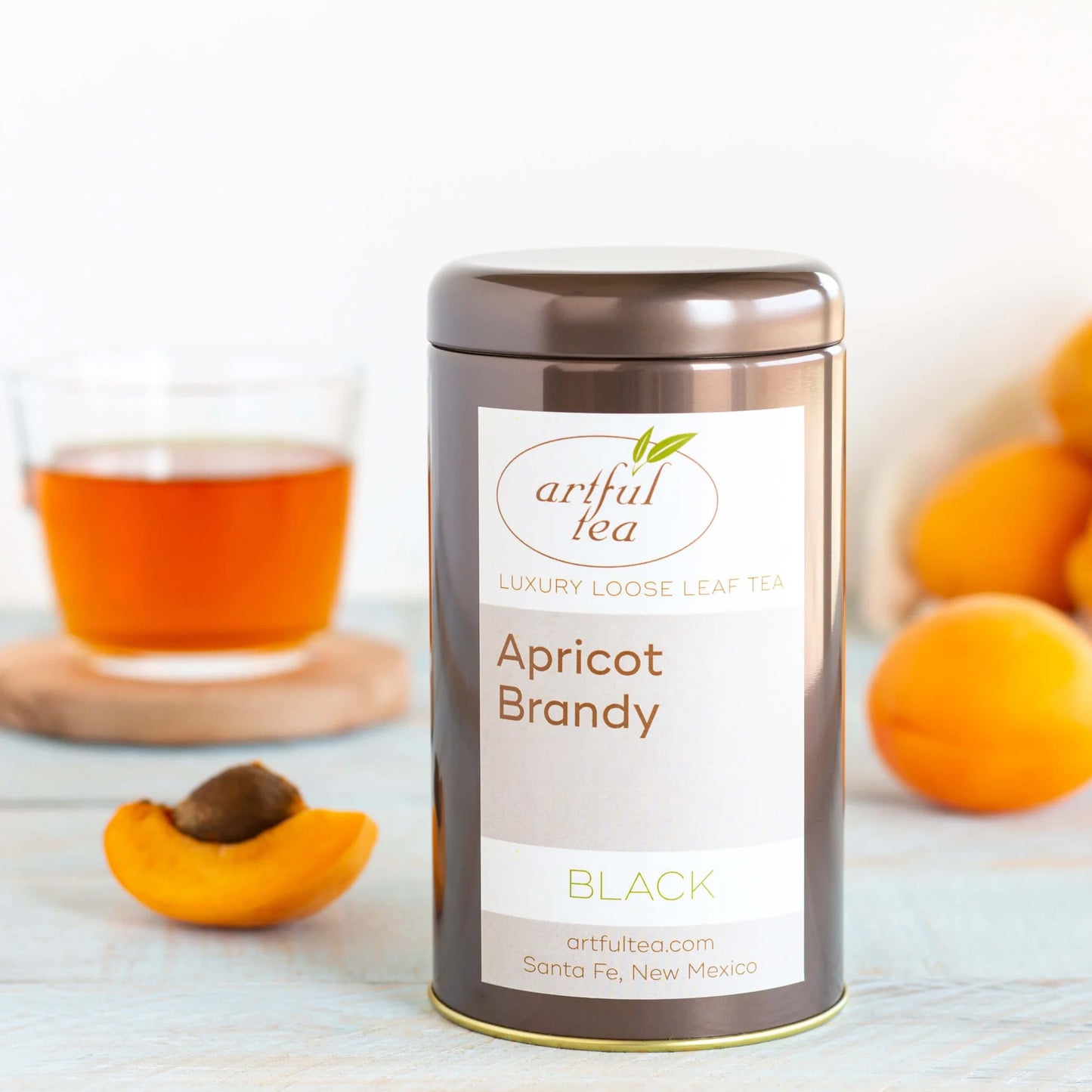 Apricot Brandy Black Tea shown packaged in a brown tin with apricots nearby and a clear cup of brewed tea on a wooden coaster.